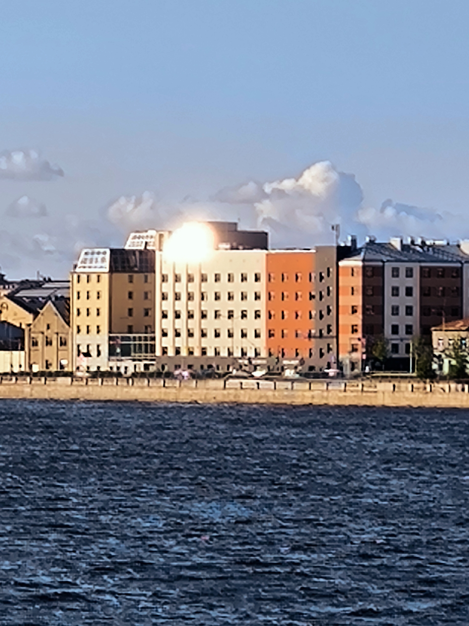 A cityscape shot shows a row of multi-story buildings, likely apartments or offices, situated along a waterfront. The buildings display a mix of architectural styles and colors, including beige, orange, and grey, with numerous windows visible. In the background, a bright light source, possibly the sun, is reflecting intensely off one of the buildings, creating a strong glare. Above the buildings, a blue sky is dotted with white, fluffy clouds. In the foreground, a dark blue body of water with visible ripples fills the bottom portion of the frame. The scene appears to be located in Riga, Latvia, as the architecture and waterfront setting are characteristic of the city, particularly along the Daugava River. No people or specific actions are discernible in the image.