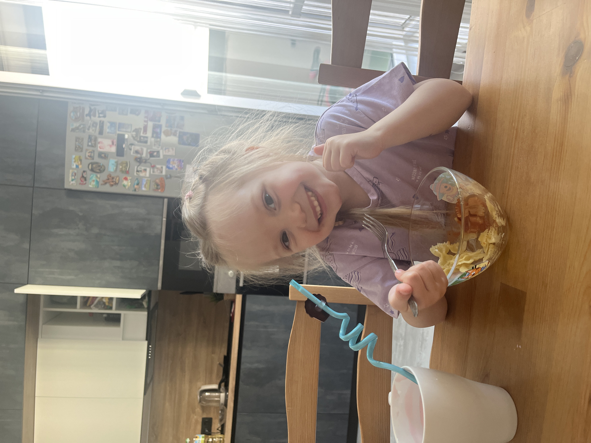A young girl with blonde hair tied up with pink bows sits at a wooden table in a modern kitchen. She is wearing a purple t-shirt and is smiling at the camera, giving a thumbs up with her right hand. In her left hand, she holds a fork, which she is using to eat pasta from a glass bowl. A white mug with a pink beverage and a blue swirly straw is to her left. The kitchen features dark grey cabinets with a wooden countertop and backsplash, and white upper cabinets. A refrigerator covered in magnets is visible in the background, as is a window with sheer curtains. The scene is lit by natural light. There are no visible identifying landmarks that would indicate the location in Chelyabinsk, Russia.