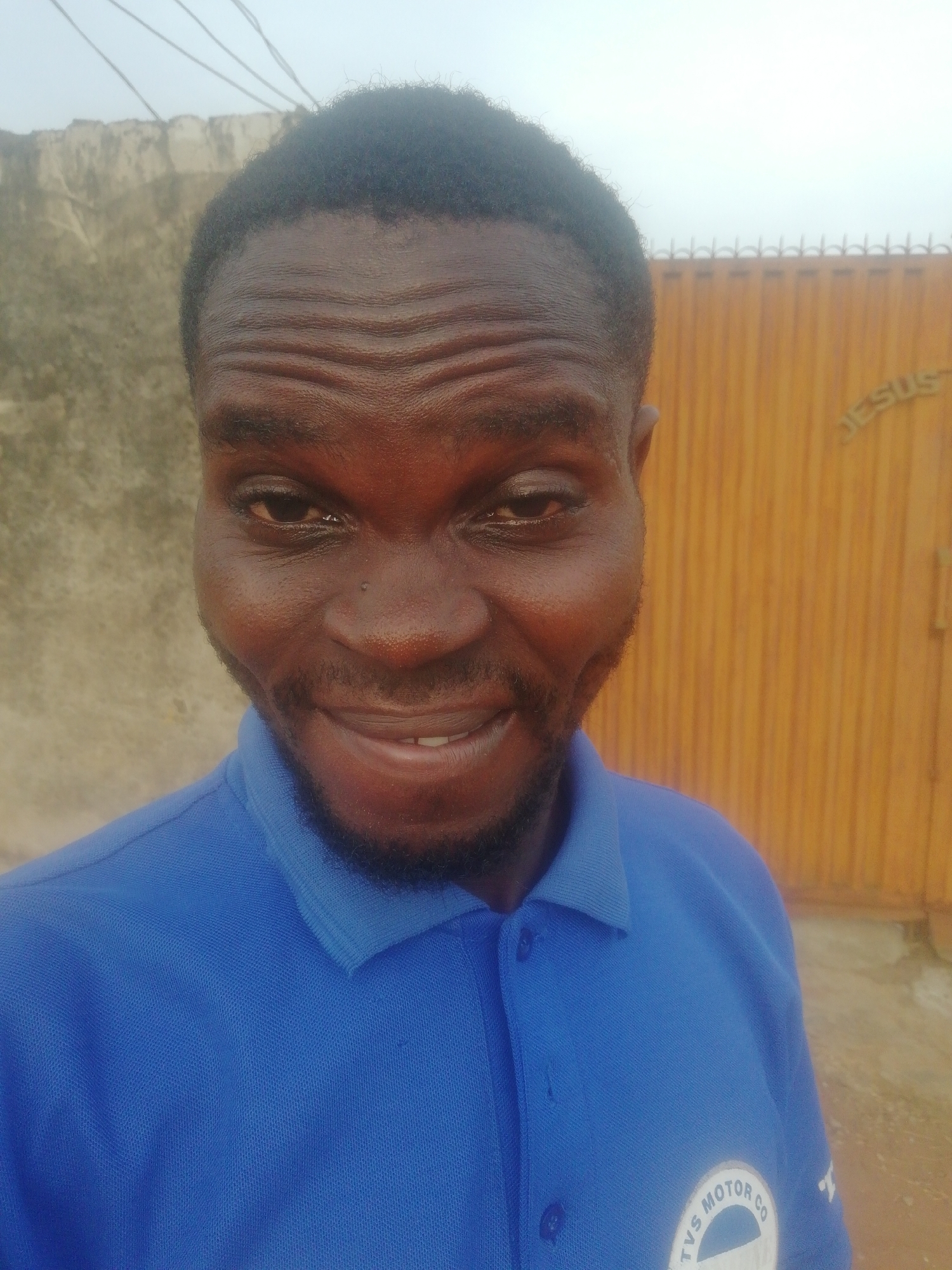 A close-up shot captures a smiling man with dark skin, wearing a blue polo shirt. He has short, dark hair and a neatly trimmed beard. Behind him, there is a weathered concrete wall and a yellow-orange gate with the word "JESUS" faintly visible. The city and specific location are not discernible from the image.