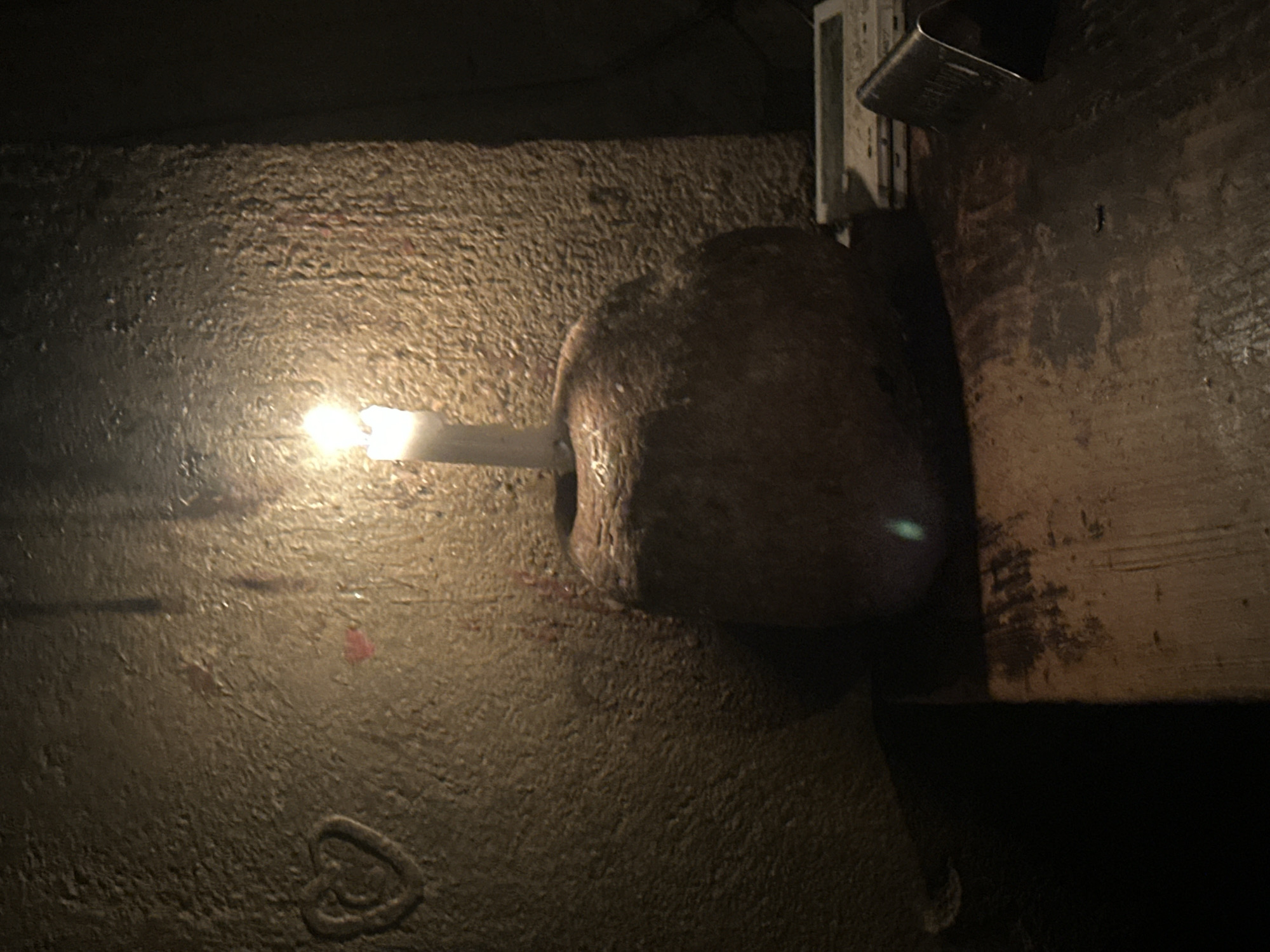 The image captures a dimly lit indoor scene, illuminated primarily by a single, lit candle. The candle is embedded into a dark, textured object, possibly a stone or a rough piece of wood, which rests on a granular, uneven surface that resembles concrete or plaster. The candle's warm, yellow-orange flame casts light across these surfaces, emphasizing their rough textures.
To the right, a darker, possibly wooden, surface is visible, extending vertically. Attached to this dark surface is a small, light-colored fixture with metallic components, which appears to be part of an electrical unit or a small bracket.
In the lower left area of the illuminated granular surface, a faint, heart-shaped outline is discernible, alongside some subtle reddish marks. The overall setting conveys a sense of quietude and intimacy, likely due to the low light conditions, suggesting it is either nighttime or a very dark interior space. No individuals or visible text are present in the frame.