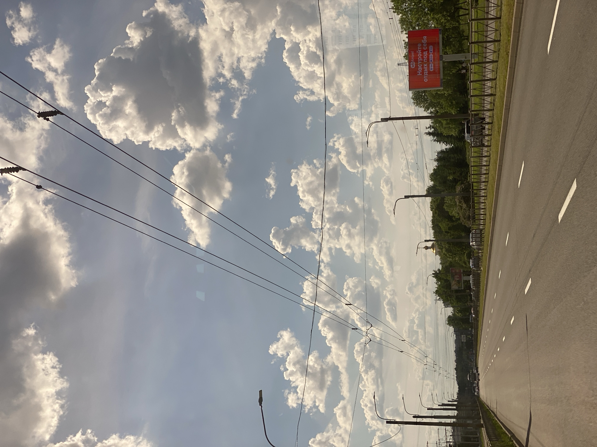 The image presents a daytime scene along a road in Saint Petersburg, Russia, under a bright, partly cloudy sky. The perspective suggests it was taken from inside a moving vehicle.
On the right side of the image, a multi-lane asphalt road or highway stretches into the distance, marked with clear white lane lines. A green median or shoulder, bordered by a metal fence, separates the road from a dense line of trees. Beyond the trees, a series of utility poles with complex overhead wires (possibly for trolleybuses or power lines) run parallel to the road. Streetlights are also mounted on these poles, extending into the background.
The sky, occupying the upper and left portions of the image, is a bright blue, filled with numerous large, fluffy white cumulus clouds. The distinct shapes and shadows of the clouds indicate bright sunlight, suggesting the time of day is late morning or afternoon.
A prominent red billboard is visible above the treeline. It displays the "SO travel" logo and the Russian text "НАСТРОЙТЕ ОТДЫХ ПОД СЕБЯ," which translates to "CUSTOMIZE YOUR VACATION." Below this, small icons are visible, likely representing various travel options. In the far distance, above the trees, a golden dome, potentially part of a church or government building, can be seen. Faint reflections of buildings suggest the photograph was taken through a window, possibly of a car or bus. The scene depicts an ordinary urban or suburban landscape on a clear, sunny day.