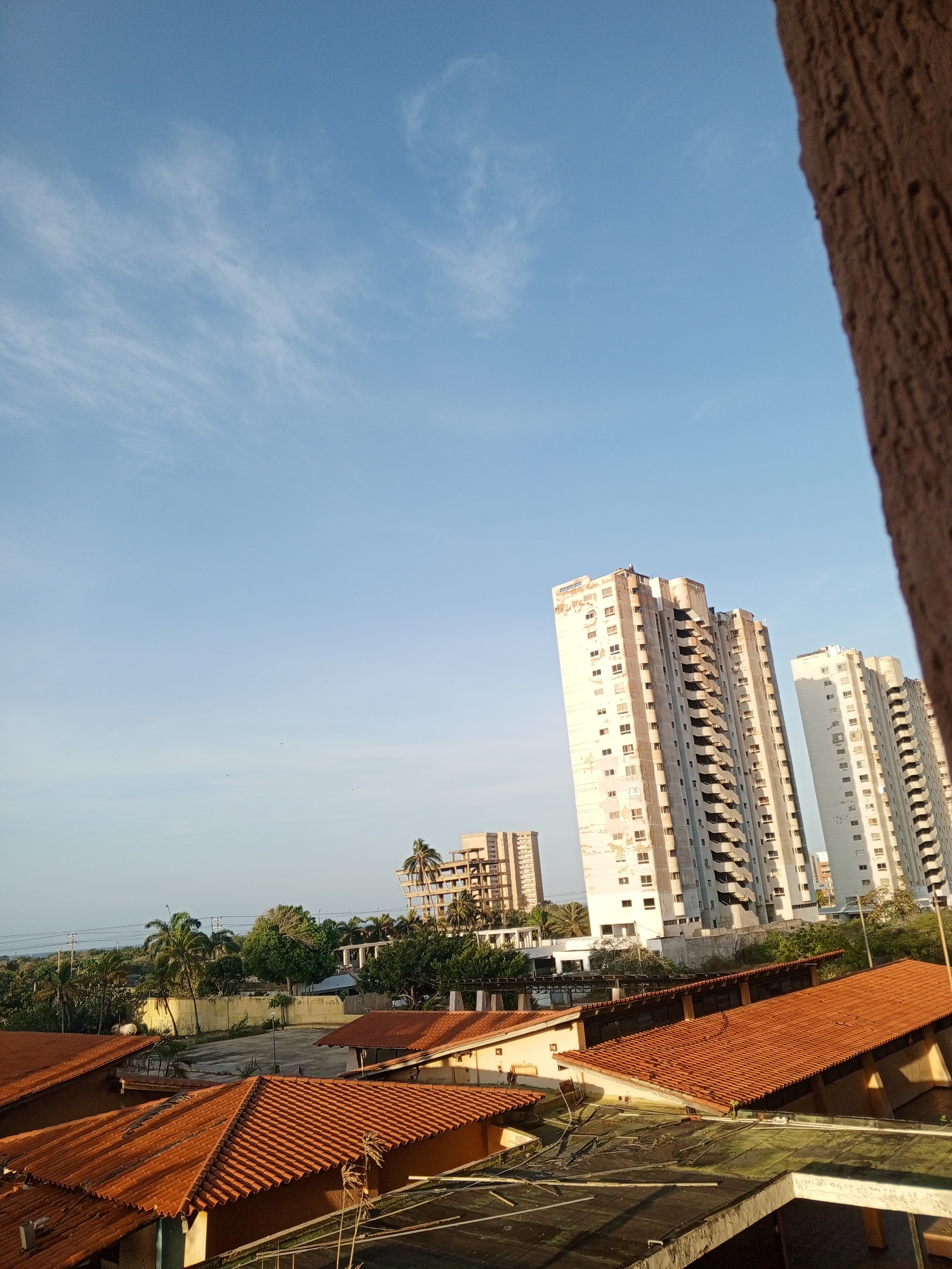The image shows a daytime view of Porlamar, Venezuela. The sky is blue with thin clouds. In the midground are several tall, rectangular buildings with many windows, some appearing aged or under construction. Below these are a number of smaller buildings with orange tile roofs. There is greenery, including palm trees, visible around the smaller buildings. In the extreme foreground, on the right side, is a textured, brown vertical structure, likely part of the building the photo was taken from.