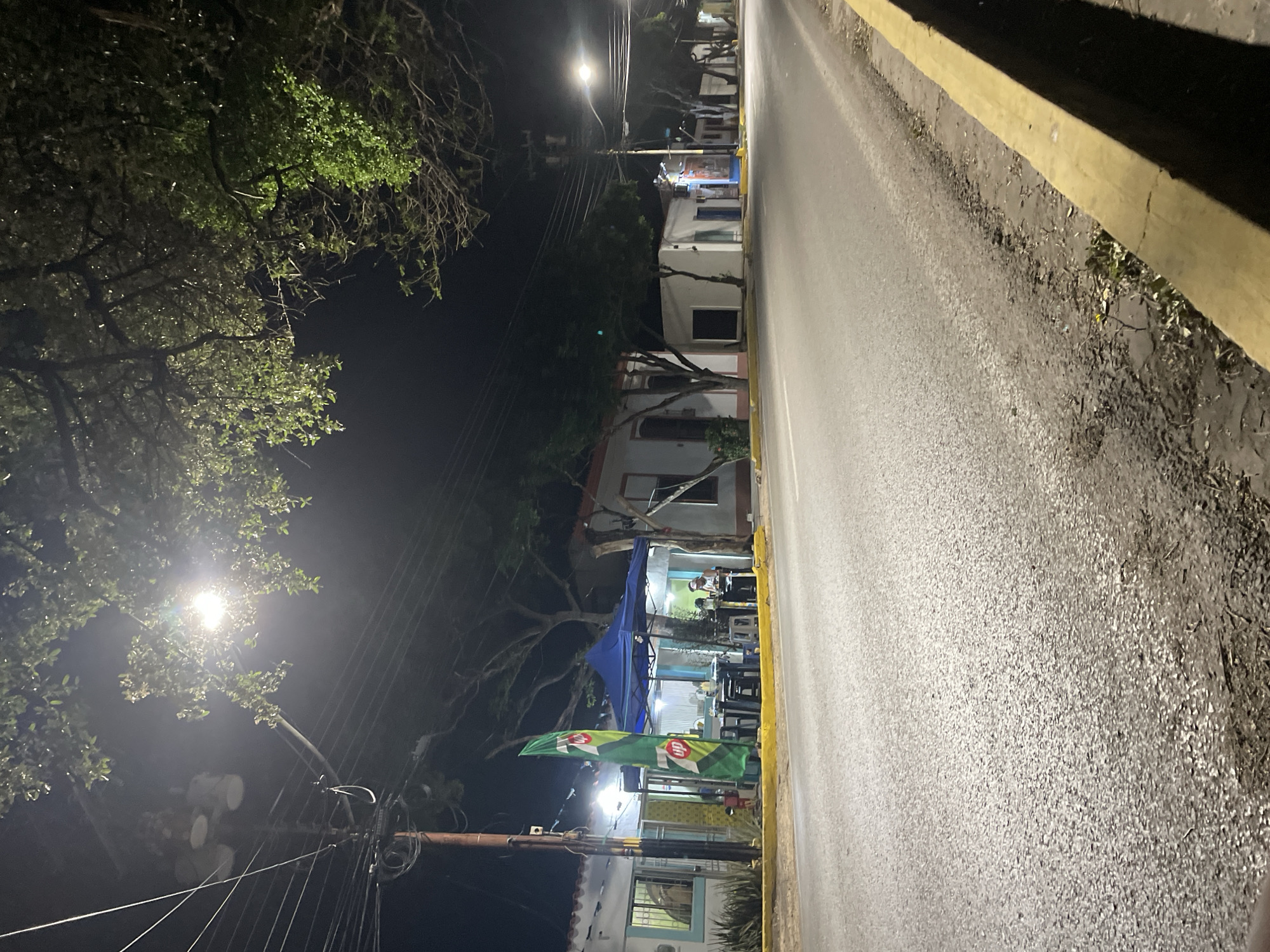 This nocturnal image, taken in Tacarigua, Venezuela, captures a street scene featuring an outdoor eatery alongside a paved road. The perspective is slightly angled, looking down the street with a prominent yellow curb on the right.
On the left side of the street, various buildings, primarily one or two stories high, are visible. Overhead, the lush canopies of trees are illuminated by the glow of streetlights, creating dappled patterns of light and shadow. A complex network of utility poles and wires crisscrosses the frame, common in many urban and semi-urban areas.
Beneath a large blue canopy, situated near one of the buildings, several individuals are seen seated at tables and chairs. They appear to be dining or socializing in what seems to be a casual, open-air restaurant or eatery. The area under the canopy is brightly lit, contrasting with the dark surroundings. Two green banners are displayed, one clearly featuring the "7UP" logo in white and red, indicating a soft drink brand.
The asphalt road, which appears dry, stretches into the distance, reflecting the light from multiple streetlights that punctuate the night sky. The overall ambiance suggests a typical evening gathering at a local establishment, characterized by a relaxed and social atmosphere.