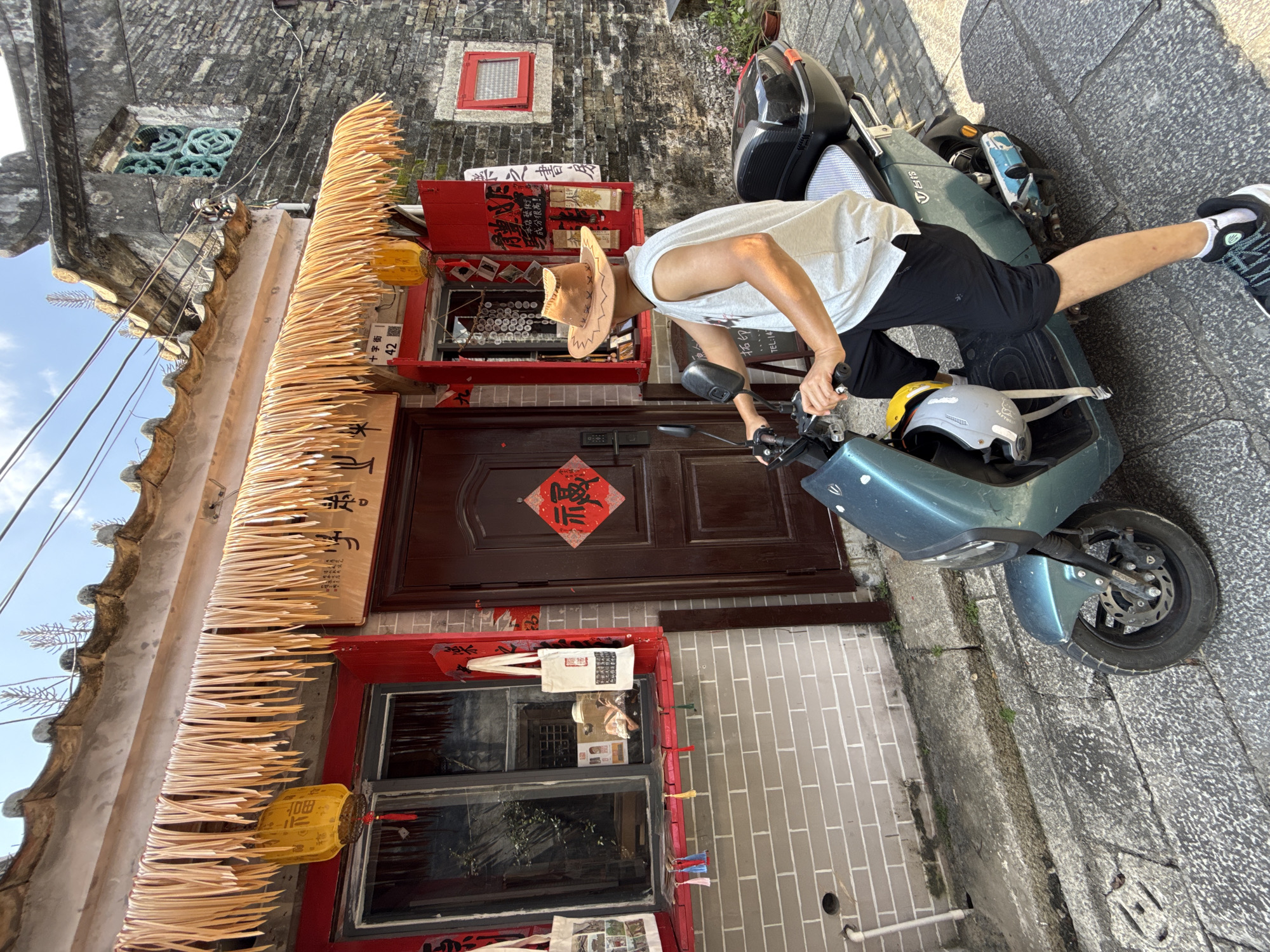 In the city of Longgang District, China, on a cobblestone street, a person wearing a cowboy hat, a white sleeveless shirt, and black shorts sits on a teal electric scooter. The scooter is parked in front of a building with a red-trimmed storefront and a thatched awning adorned with yellow lanterns. Chinese calligraphy and red decorative paper with Chinese characters are visible on the building. The building's facade is constructed with brickwork, and an old-fashioned window with decorative metalwork is visible on an adjacent structure. The scene appears to be an outdoor marketplace or a historic street.