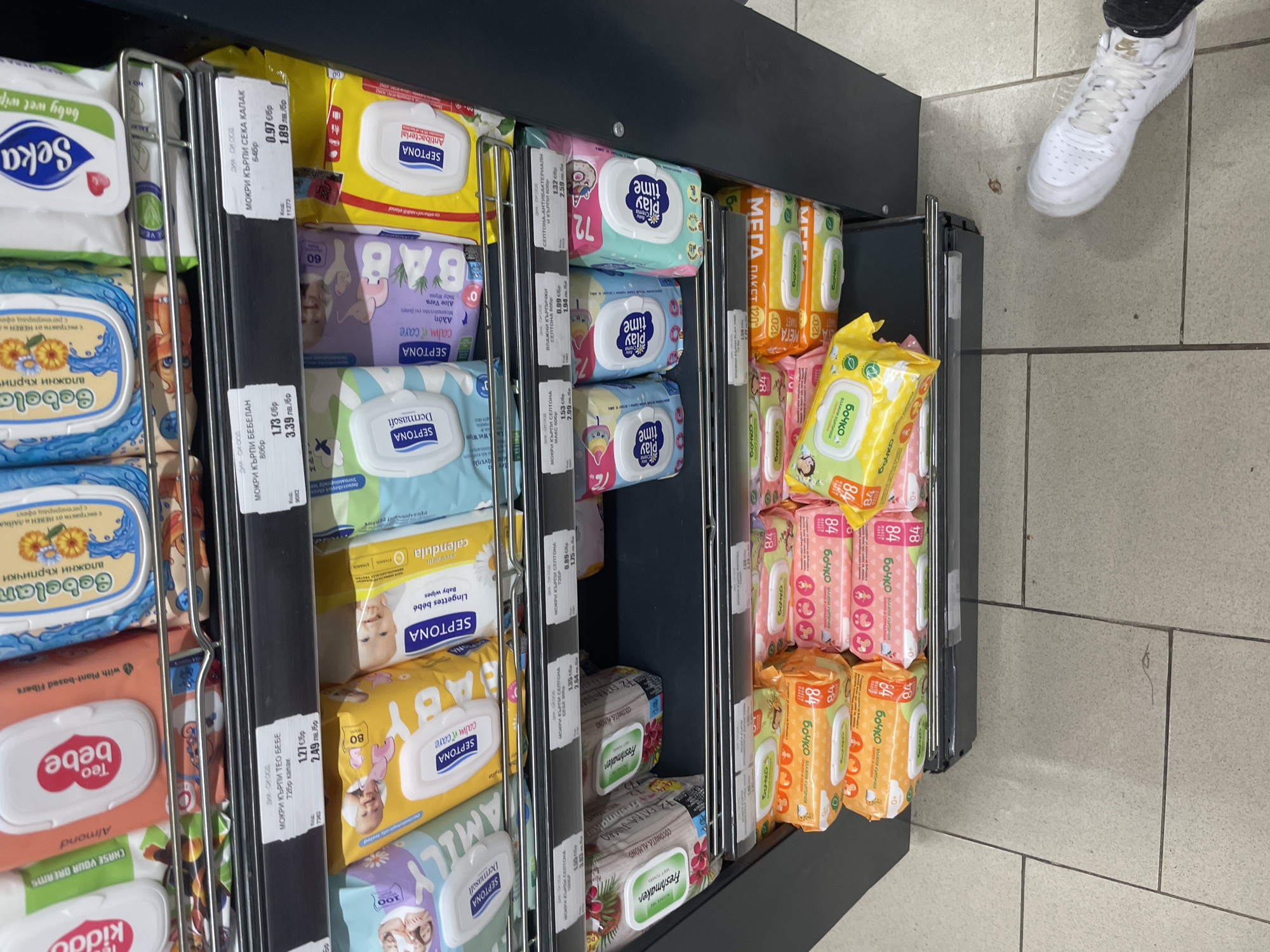 The image captures a store aisle in Ihtiman, Bulgaria, specifically showcasing a metal display rack stocked with numerous brands and varieties of wet wipes and baby wipes. The rack is dark grey with vertical metal dividers, organizing the products into neat rows and stacks.
Visible brands include Septona, Bebelan, Teo Bebe, Play Time, Freshmaker, Бочко (Bochko), Seka, and Kiddo. The products offer diverse features: Septona has antibacterial, calm & care (with Aloe Vera), Dermasoft, and calendula baby wipes. Bebelan wipes highlight extracts of calendula and chamomile. Teo Bebe wipes are almond-scented, and Freshmaker offers coconut and almond varieties. Бочко (Bochko) wet wipes are prominently displayed in multiple colors, often in 84-count packs, with some "Mega Pack" options containing 120 wipes. Play Time wipes come in colorful 72-count packages. Other common pack sizes indicated are 50, 60, 80, and 100 wipes.
Price labels in Bulgarian (лв.) are attached to the shelves. Examples of prices per pack include: Seka wet wipes (64 pcs) for 1.89 лв; Bebelan wet wipes (80 pcs) for 3.39 лв; Teo Bebe wet wipes (72 pcs) for 2.49 лв; Septona Antibacterial wet wipes (60 pcs) for 2.59 лв; and Septona MAX wet wipes (50 pcs) for 2.99 лв.
In the upper right, a person's foot, wearing a white Nike sneaker, is visible on the light grey tiled floor, indicating someone is standing near the display. The setting is an indoor retail environment with artificial lighting. No specific activity beyond browsing or standing is discernible, and no emotions are evident.