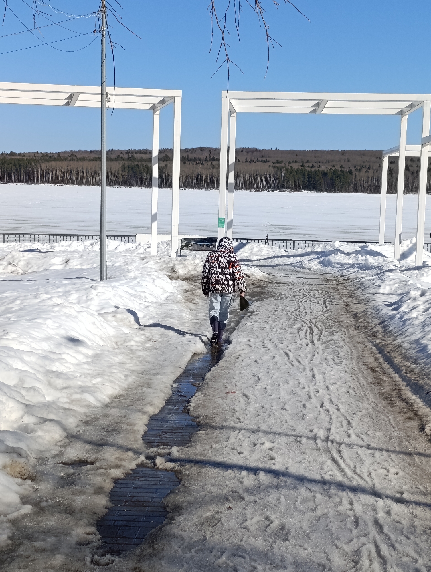 An individual walks away from the camera along a partially cleared paved pathway on the city embankment in Chaikovsky, Russia. The pathway, lined with melting snow and slush, reveals sections of dark paving stones and puddles of water reflecting the sky. The person, viewed from behind, wears a multi-patterned jacket with text, light-colored pants, and dark boots, holding a dark object in their right hand.
To the left and right, large white rectangular arch-like structures frame the view. A slender grey metal pole with wires is positioned near the left arch, with bare tree branches visible in the upper left. Beyond the pathway and structures, a dark metal fence runs horizontally, separating the land from a vast, snow-covered frozen body of water, likely the Kama River or Votkinsk Reservoir. In the far background, across the frozen water, a dense line of trees, a mix of bare deciduous and evergreens, forms the horizon. Two tiny, distant figures are discernible on the ice near the fence. The scene is illuminated by strong sunlight under a clear, bright blue sky, indicating late winter or early spring conditions with significant snowmelt.