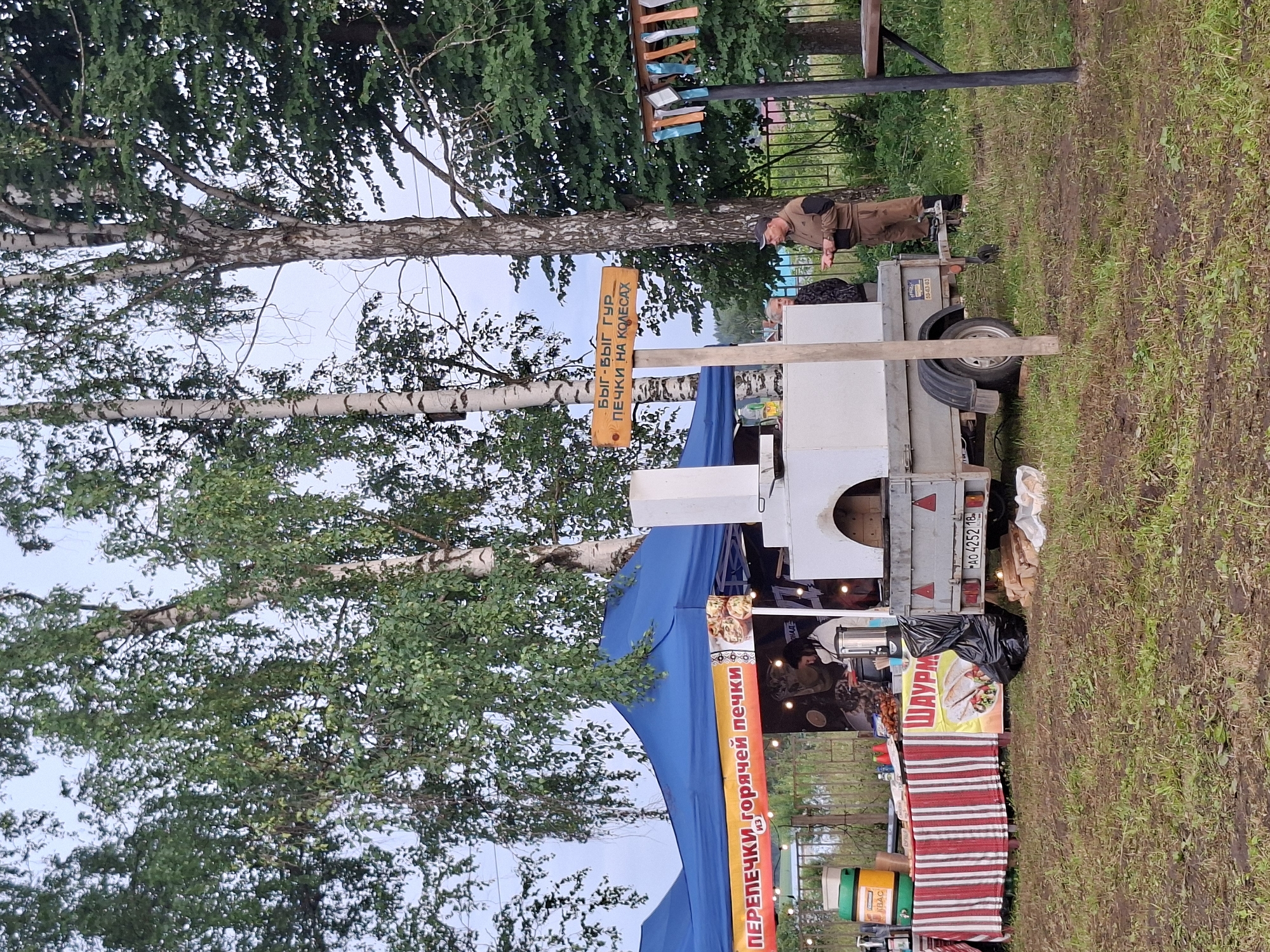 This media file depicts an outdoor scene in what appears to be a rural or park-like setting, likely in Старые Быги, Russia. The primary subjects are a food stand and two individuals. The food stand is a mobile unit, resembling a trailer, with a blue canopy. It is decorated with signs advertising food items. One prominent sign, in red and yellow, reads "ПЕРЕПЕЧКИ горячей печки" (Perepechki of hot oven) and another in yellow reads "ШАУРМ" (Shawarma). Above the stand, a wooden sign on a birch tree reads "БЫГ-ВЫГ ГУР ПЕЧКИ НА КОЛЕСАХ" (Byg-Byg Gur Ovens on Wheels), indicating the name of the food business.
One person, dressed in work clothes, stands near the trailer, gesturing with their hands. Another person is visible behind the food counter, possibly preparing food or serving customers. The ground is covered in green grass, with some muddy patches, suggesting it might have rained recently or the ground is damp. Several trees, including birch trees with their distinctive white bark, are present, with lush green foliage, indicating it is likely spring or summer.
A license plate on the trailer reads "PAO 4252 18". There are also various containers and equipment associated with a food vending operation visible, such as beverage dispensers. The overall atmosphere seems casual and functional, typical of an outdoor food vendor. The lighting suggests it is daytime, possibly late afternoon or early evening, given the diffused light and the presence of shadows.