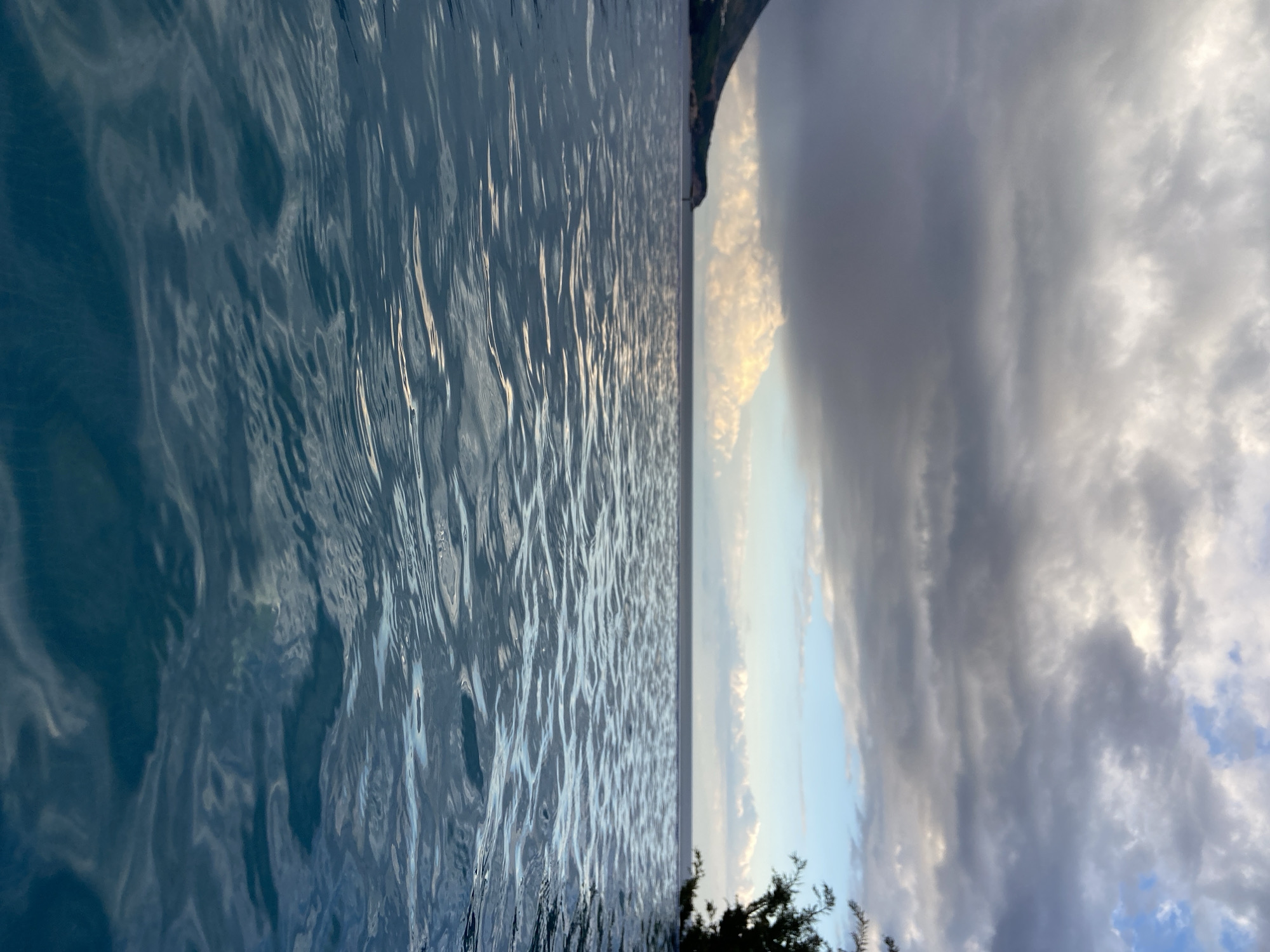 The image, oriented horizontally, captures a tranquil waterscape at what appears to be either sunrise or sunset, likely from an infinity pool or a calm body of water in Boreti, Montenegro.
The foreground and left side of the frame are dominated by the deep blue-grey surface of the water, which is gently ruffled by subtle currents. Sunlight reflects vividly off the ripples, creating shimmering streaks and intricate patterns of light and shadow across the water's surface.
A sharp, clear horizon line separates the water from the sky. Above the horizon, the sky presents a compelling contrast. Towards the left and center, there are patches of soft blue sky interspersed with luminous, golden-hued clouds, indicating the warm glow of either dawn or dusk. Moving further right and upwards, the sky transitions to heavier, dark grey clouds, suggesting either the presence of more significant cloud cover or the deepening shades of twilight.
On the right side of the horizon, a dark, indistinct landmass, likely a distant mountain or coastline, is visible, partially silhouetted against the brighter sky. In the bottom right corner, a small fragment of dark green foliage, possibly from a tree or bush, adds a touch of nature to the immediate surroundings.
The scene conveys a sense of peaceful serenity combined with the dramatic beauty of a transitional sky. There is no visible text in the image.
