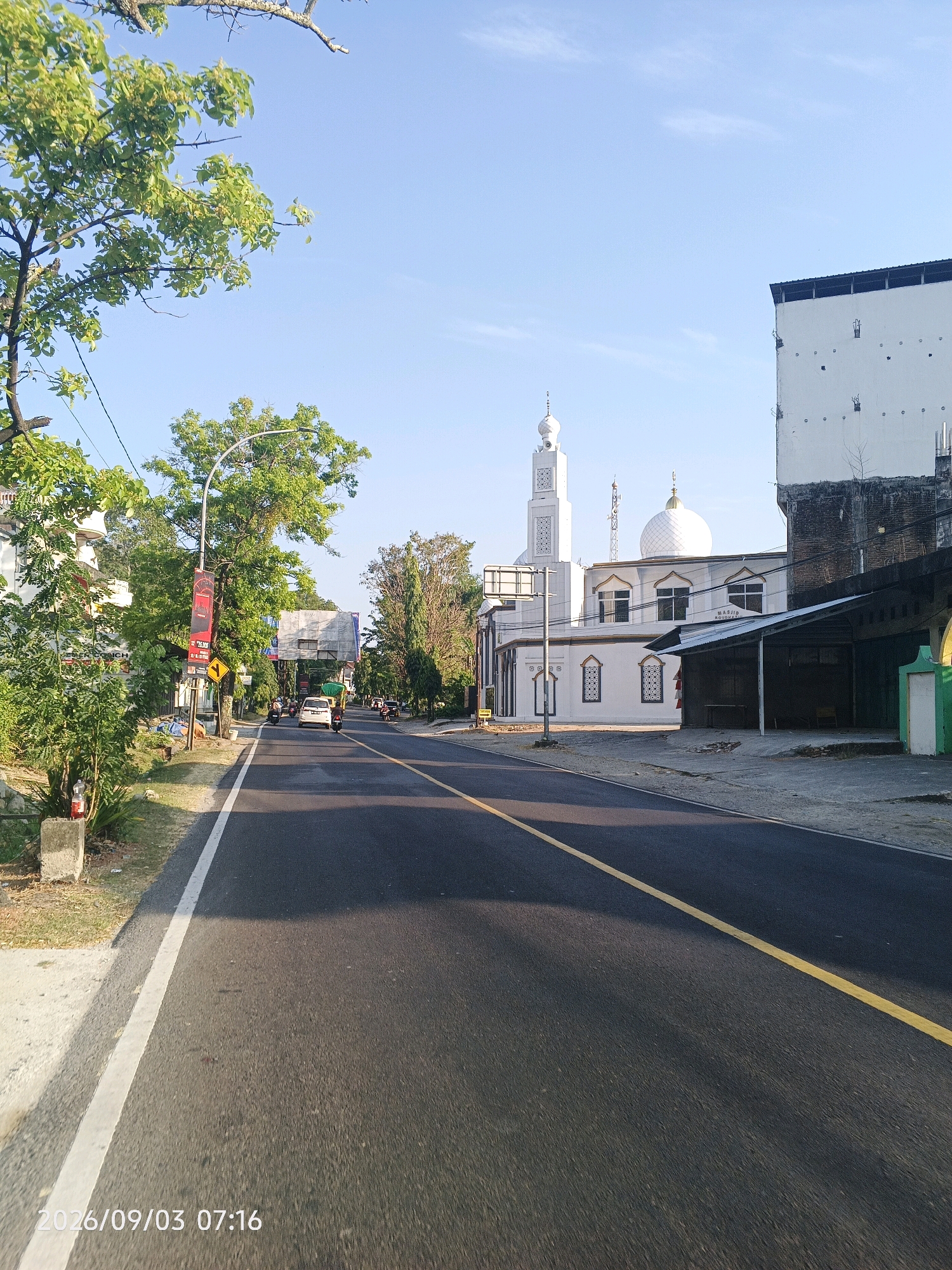 The image captures a street scene in Pare-Pare, Indonesia, on September 3, 2026, at 7:16 AM. The perspective is from the road, looking forward. On the right side of the road, a mosque is prominently visible with a white dome and a tall minaret. Adjacent to the mosque, a white building with a dark roof is under construction or renovation, with exposed brickwork on its facade. A narrow, shaded awning runs along the front of a low structure next to the mosque.
To the left of the road, trees with green foliage line the sidewalk. A traffic light and several street signs are visible, along with a red banner. A white car is driving on the road, and further down, several motorcycles and a white van are also present. The road itself is asphalt with a distinct yellow line marking the center. The sky is clear and blue, indicating a sunny morning. The overall impression is a quiet street in an Indonesian city, with a mix of residential and religious buildings, and moderate morning traffic.
