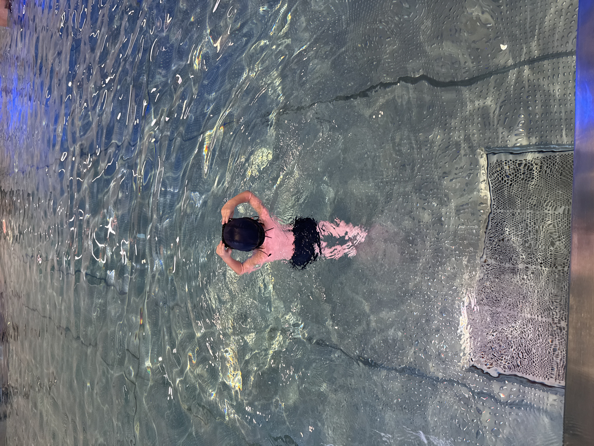 A person is standing in a swimming pool. The person is wearing a black swimming cap and dark swim trunks. They are holding their hands up to the back of their head. The water is clear and reflects the light from above, creating rippling patterns. There is a textured surface visible at the bottom of the pool in the foreground. The lighting suggests an indoor pool environment. This scene does not provide any information that would allow for its localization to Sovkhoza imeni Lenina, Russia.