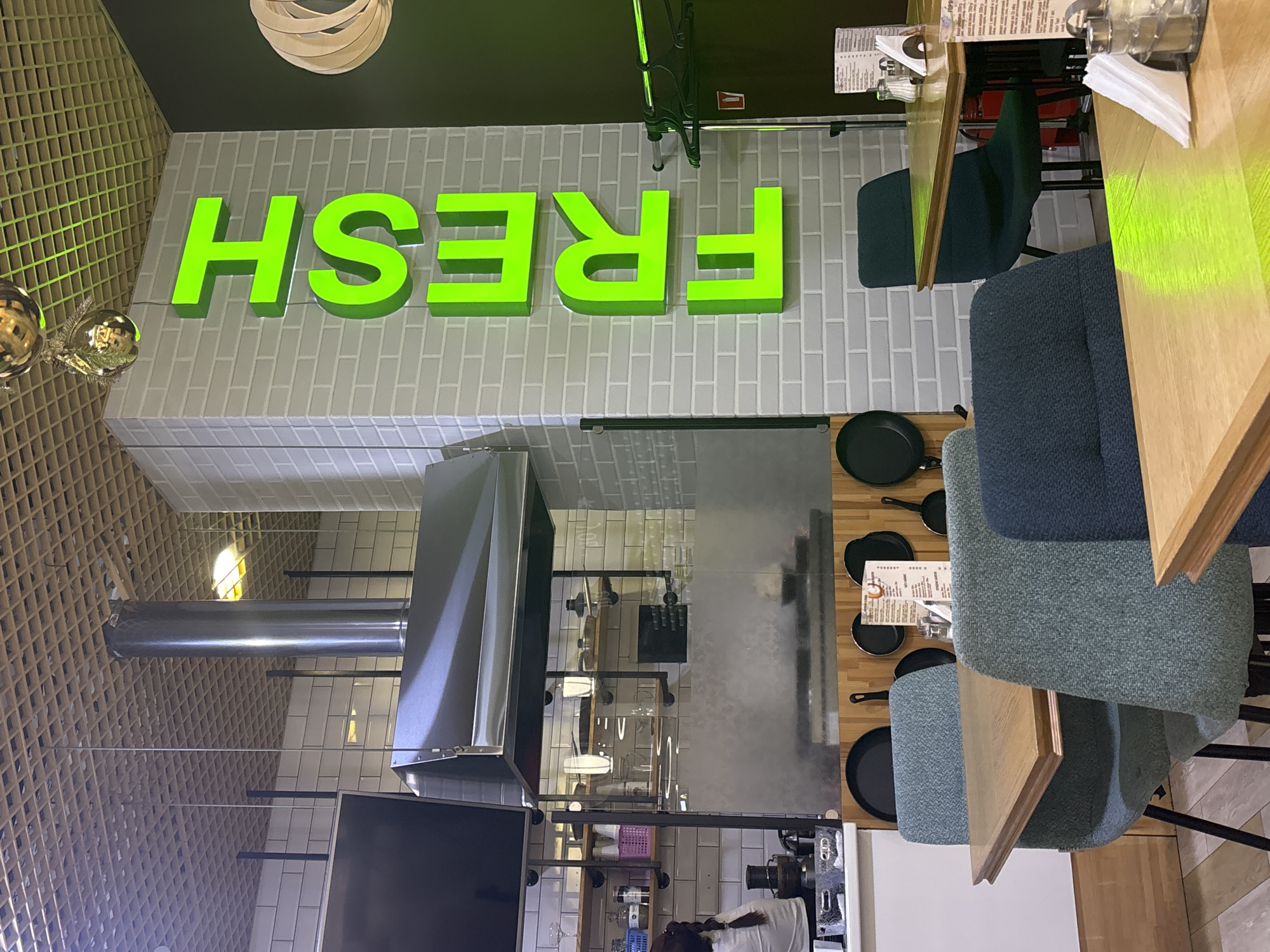 The image shows a view of a restaurant's interior. Large, bright green 3D letters spelling "FRESH" are mounted on a light grey brick wall. To the left, decorative golden spherical ornaments hang near a wooden lattice wall.
The scene also includes restaurant seating with wooden tables and chairs upholstered in blue and grey fabric. On one of the tables, a menu is visible, along with a glass of water and some napkins. Cooking equipment, including a stainless steel hood and what appears to be a stove, is present in the background, suggesting a kitchen or open-plan dining area.
One cropped image shows a person from the back, wearing a white top, with dark braided hair, working in the background. Another crop shows a glass, water bottles and a paper napkin on the table. A frying pan is visible on a table. The lighting suggests it is daytime. The text visible includes "FRESH" on the wall and some text on a menu. The location is identified as Naberezhnye Chelny, Russia.