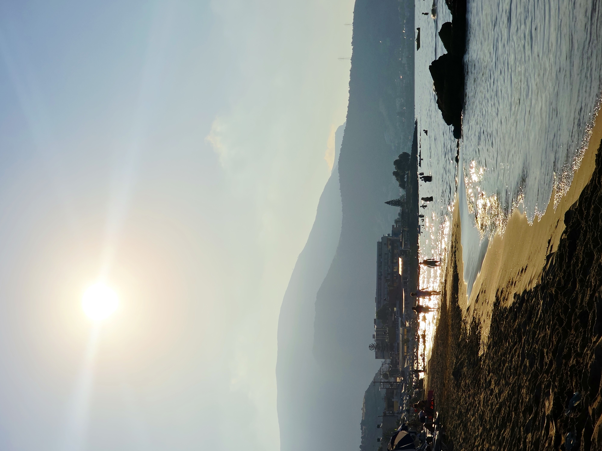 The image depicts a serene beach scene in Malia Municipal Unit, Greece, during what appears to be late afternoon or early evening. The sun is low in the sky, casting a warm, golden light across the landscape and creating a shimmering effect on the water. In the foreground, the sandy beach is textured with ripples and shadows. To the left, beach umbrellas and lounge chairs are visible, some occupied by people.

The middle ground features the shoreline where several individuals are walking into the shallow water or standing near the edge. Their silhouettes are distinct against the bright water and the sun's glare. Further out in the sea, more people are swimming or wading.

In the background, a line of buildings, likely hotels or shops, stretches along the coast, silhouetted against the soft light. Beyond the buildings, rolling hills and mountains form a gentle, hazy backdrop. The sky is a pale blue, with the bright sun as the dominant light source, creating lens flare effects. A few scattered clouds are present. The sea is calm, with gentle waves lapping the shore. A small boat is visible in the distance. The overall atmosphere is tranquil and summery.