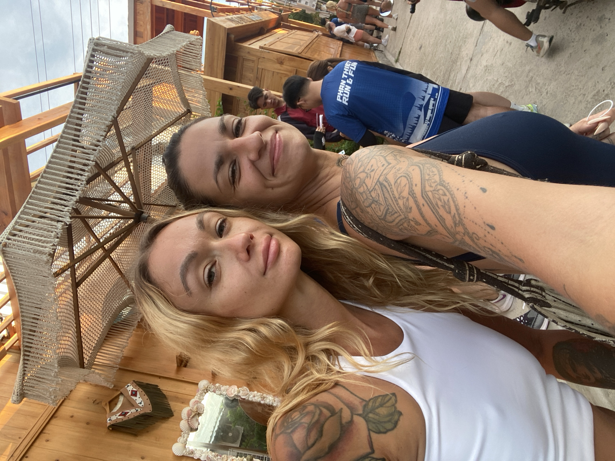 Two women are taking a selfie outdoors in what appears to be a street market or outdoor dining area. The woman on the left has blonde hair and is wearing a white tank top with a large rose tattoo on her left shoulder. The woman on the right has dark hair and is wearing a dark blue top, with visible tattoos on her arms. Behind them, wooden structures suggest a commercial or entertainment venue. In the background, several other individuals are visible, including a man wearing a blue shirt with "PHAN THIET RUN & FUN" printed on it. The environment suggests a warm, possibly tropical climate. The scene is likely located in Phu Thuy Ward, Phan Thiet City, Binh Thuan Province, Vietnam, as indicated by the text on one of the shirts.