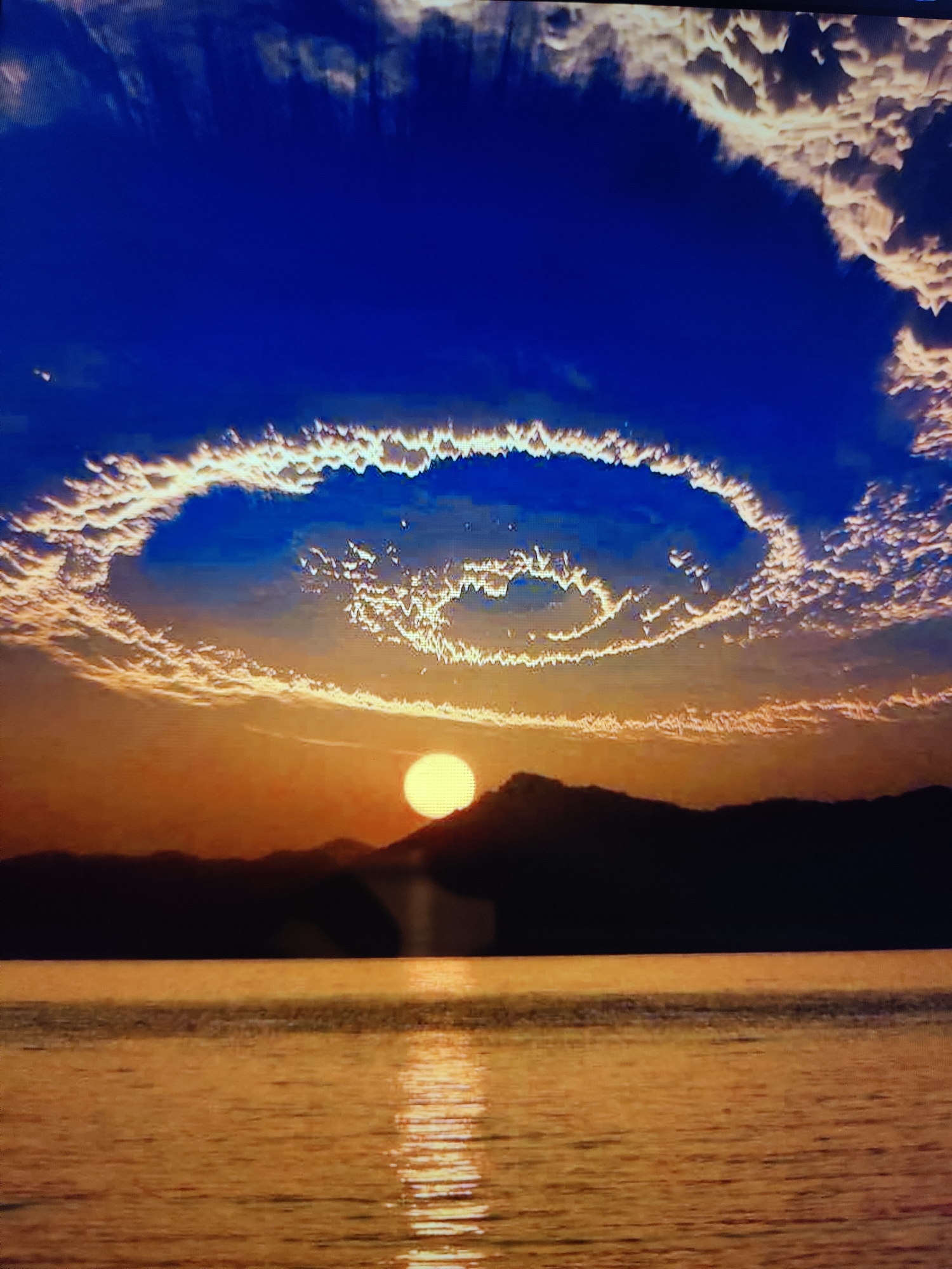 The image depicts a dramatic sunset over the Caspian Sea, likely viewed from Baku, Azerbaijan. The sky is a deep, vibrant blue, with a ring of luminous, wispy clouds forming a halo around a bright, yellow sun. The sun is partially obscured by a dark, silhouetted mountain range in the distance. The Caspian Sea in the foreground reflects the warm, golden hues of the sunset, with shimmering light stretching across its surface. The overall scene is one of natural beauty and tranquility.