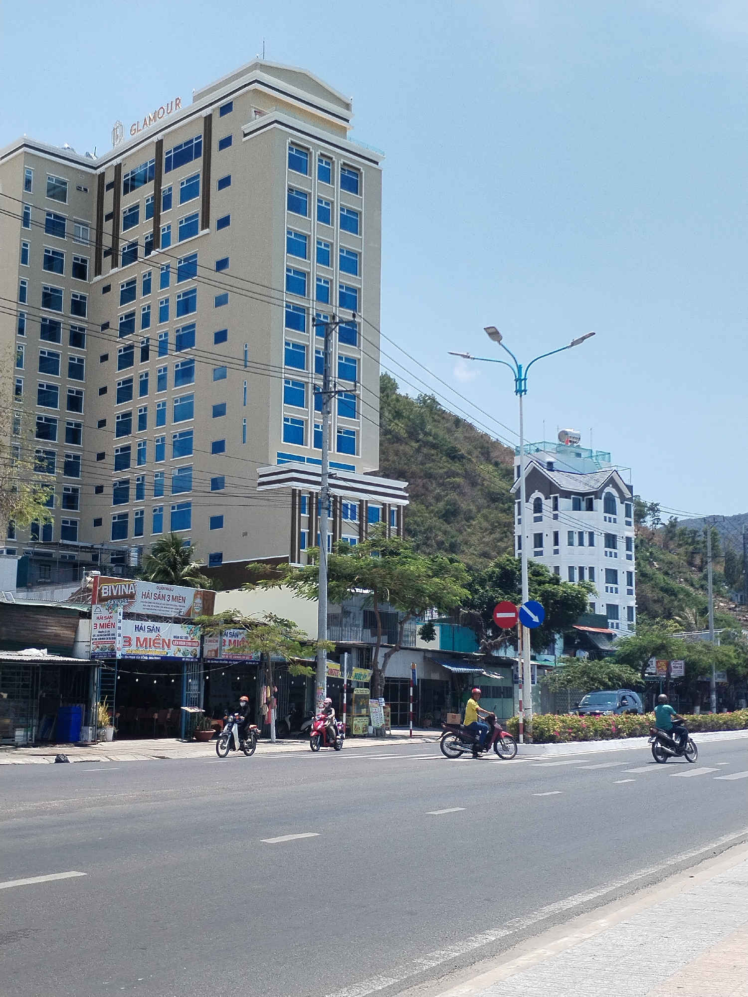 The image shows a street scene in Bac Nha Trang Ward, Vietnam. A tall, multi-story beige building with blue windows, labeled "GLAMOUR" at the top, dominates the left side of the frame. Below this building are several storefronts with signs, including one that says "BIVINA HẢI SẢN 3 MIỀN". The street in the foreground is a grey asphalt road with white lane markings. Three motorcyclists are visible, traveling along the road. In the background, another multi-story white building stands against a backdrop of green hills. A blue street light and a traffic sign are also present. The sky is clear and blue.