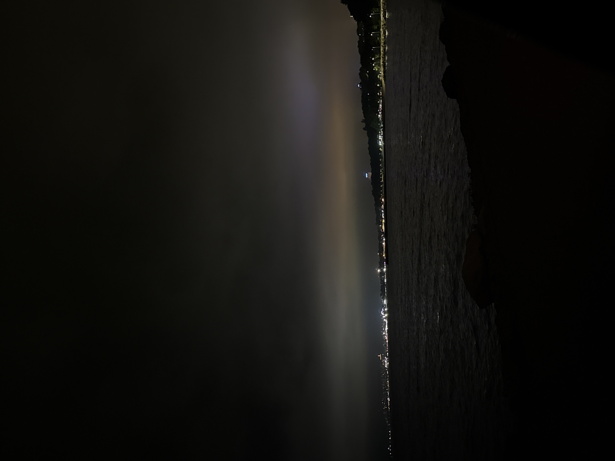 A forensic examination of the image reveals a nighttime coastal landscape of the city of Varna, Bulgaria, viewed from a dark, unpopulated shoreline.
The foreground is dominated by the silhouette of dark rocks and a section of a breakwater or beach, occupying the lower quarter of the frame. Immediately beyond, a vast expanse of dark, subtly rippling water, identified as the Black Sea, fills the midground. The water reflects minimal light, appearing as a deep, somber tone.
Along the horizon, the illuminated coastline of Varna stretches from left to right. Clusters of artificial lights, predominantly white, yellow, and orange, mark the urban development. A tall, slender structure with a distinct red light at its zenith is visible towards the center-left of the city lights. Further to the right, the land rises into a densely vegetated hill, likely part of the Sea Garden, with a visible road winding along its base, illuminated by equidistant streetlights. On the crest of this hill, a prominent, well-lit, white-domed structure, identifiable as the Varna Observatory/Planetarium, stands out against the darkness.
The sky is heavily overcast, absorbing ambient light. However, the low-hanging clouds are illuminated from below by the city's light pollution, creating a diffuse glow that transitions from a dark grey-brown near the top of the frame to warmer yellowish and purplish hues directly above the city lights.
No human figures, animals, or specific actions are discernible within the frame. The scene is static, capturing the urban glow of Varna under a cloud-covered night sky.