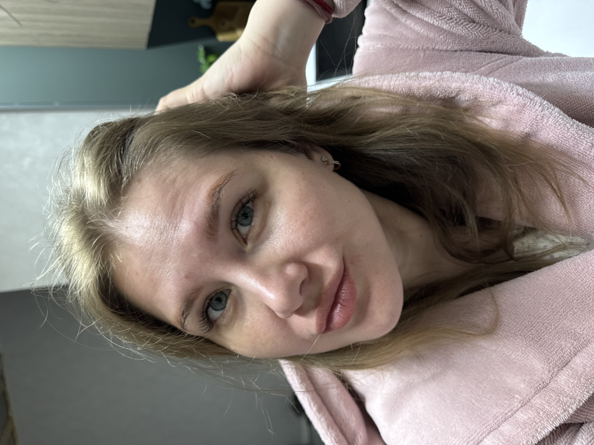 A close-up portrait shows a young woman with light brown/blonde hair and light blue-green eyes, looking directly at the camera with a gentle, serene expression, her lips slightly parted in a faint smile. She is wearing a soft, textured, light pink robe or hooded garment. Her head is tilted, and her left hand is positioned near the top of her head, lightly touching her hair. A small gold hoop earring with a heart charm is visible in her right ear, and a red elastic band is on her left wrist. The background is softly blurred but suggests an indoor domestic setting, possibly a kitchen or living area, with hints of a dark countertop, a lighter wall, and some indistinct objects like a wooden board and greenery. The overall lighting appears natural, suggesting it is daytime. The image is taken at an angle, giving it a casual, candid feel. There is no visible text in the image. The scene takes place in Gornyi (Dvurechenskoe S/P), Russia.
