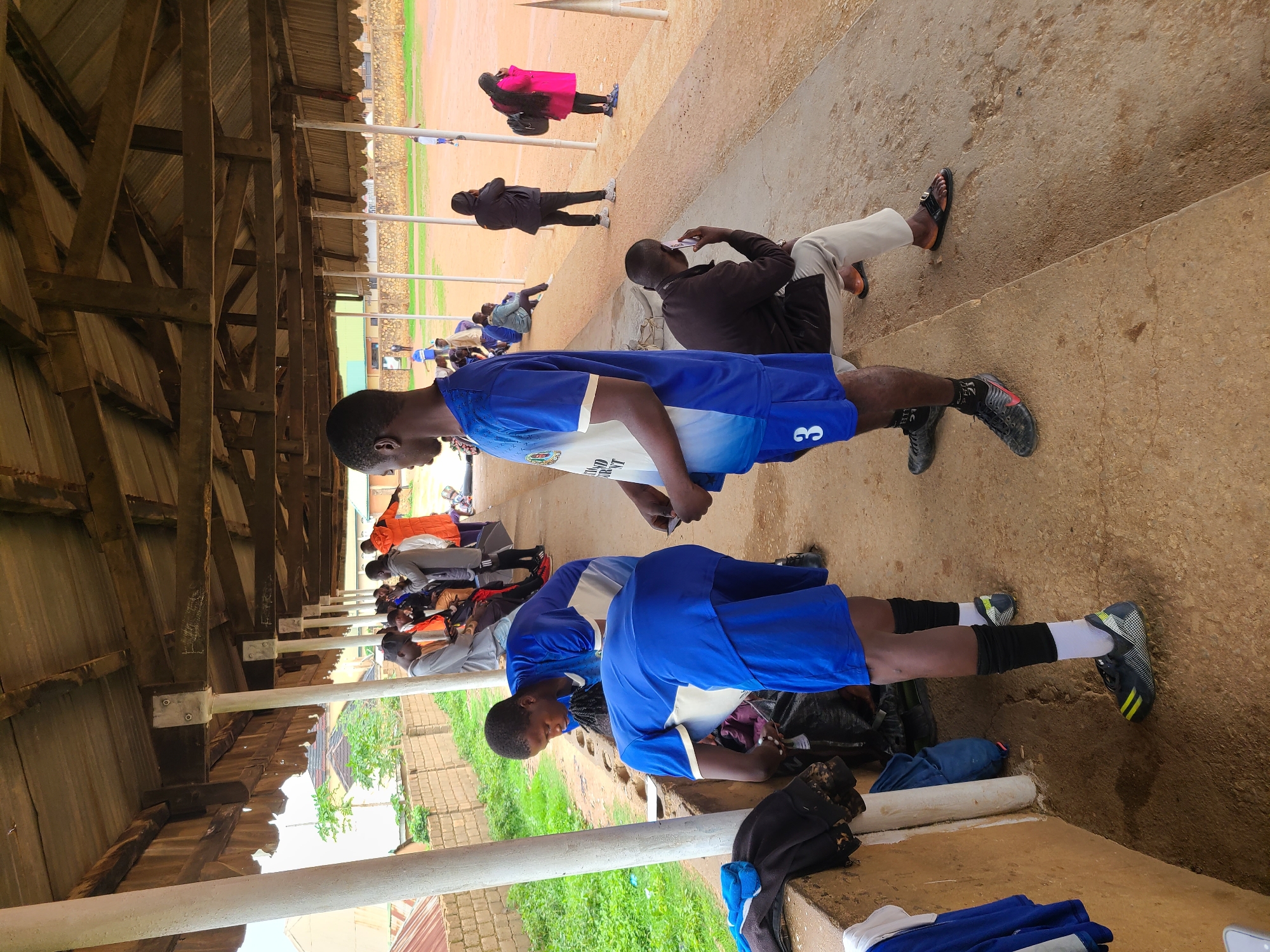 The image shows a group of people sitting and standing under a covered structure, likely a waiting area or shelter. Two people in blue and white soccer uniforms are visible in the foreground. One is standing and the other is kneeling. The standing person has the number 3 printed on their shorts. Some of the other people are sitting on a concrete platform, and one person is on a phone. Further in the background, more people are standing and walking around on a field, which appears to be a sports field with grass and dirt. A person in a pink jacket is walking in the far background. The location is Unknown, Unknown.