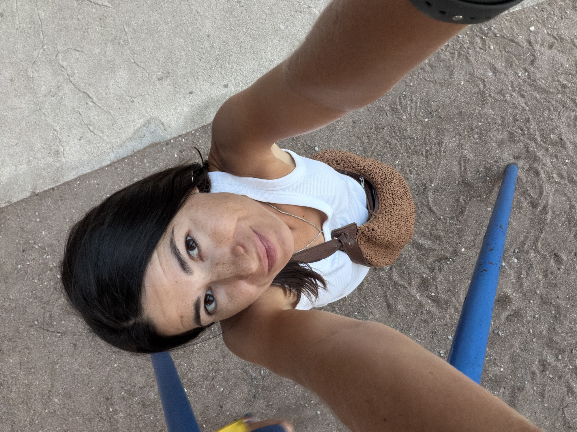 The image is a high-angle, point-of-view selfie taken by a woman outdoors, likely on a beach or near a playground in Budva, Montenegro. The woman has dark hair, brown eyes, and freckles. She is wearing a white tank top and a small, woven brown bag with a strap across her chest. In the foreground, there are blue bars, suggesting playground equipment. The ground is covered in sand with small pebbles, and there is a concrete surface in the upper right portion of the image. The lighting indicates it is daytime.