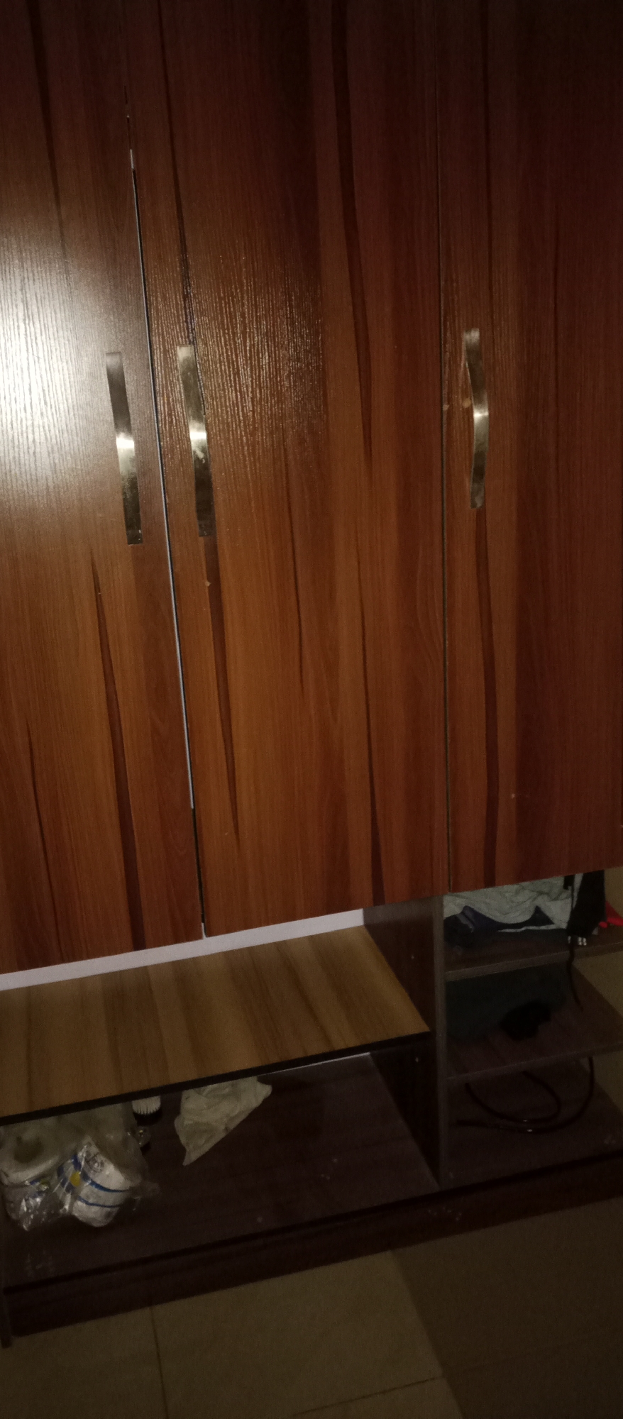 The image shows a close-up view of a wooden wardrobe with three doors. The doors have metallic handles. Below the wardrobe, there are shelves with some items on them, including what appear to be clothes and some packaged goods. The lighting suggests an indoor environment. The location is identified as Gwarinpa, Nigeria.