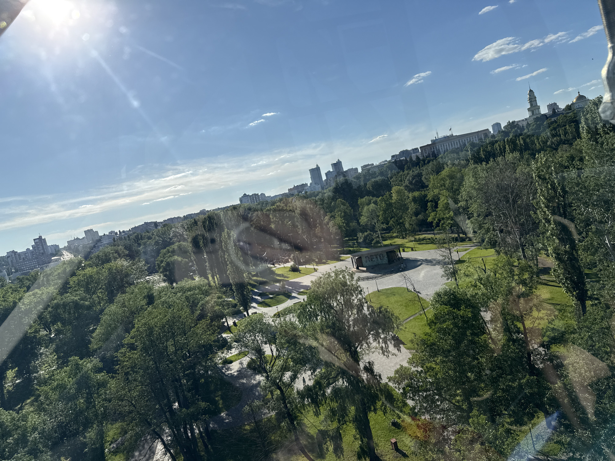The image presents an elevated view of a sunlit urban landscape in Lipetsk, Russia, captured through a reflective glass surface displaying lens flare. The central and foreground portions are dominated by an extensive green parkland, characterized by numerous mature deciduous trees and a network of paved pedestrian pathways. Scattered individuals are present within the park, with one person visible cycling on a path. A modern, rectangular building, partially visible, bears the Cyrillic inscription "ЛЕТО" (LETO, meaning "Summer") on its side, suggesting a park facility. In the background, a clear blue sky with sparse white clouds hangs above the city. Key architectural landmarks are identifiable: the Cathedral of Christ's Nativity, marked by its tall, light-colored spire, is prominent on the right. Adjacent to it, the Lipetsk State Academic Drama Theatre features a distinctive golden dome. Further to the left, a large, columned building, consistent with the Lipetsk Oblast Administration, is visible, displaying a flag. Various other multi-story buildings constitute the cityscape on the horizon.