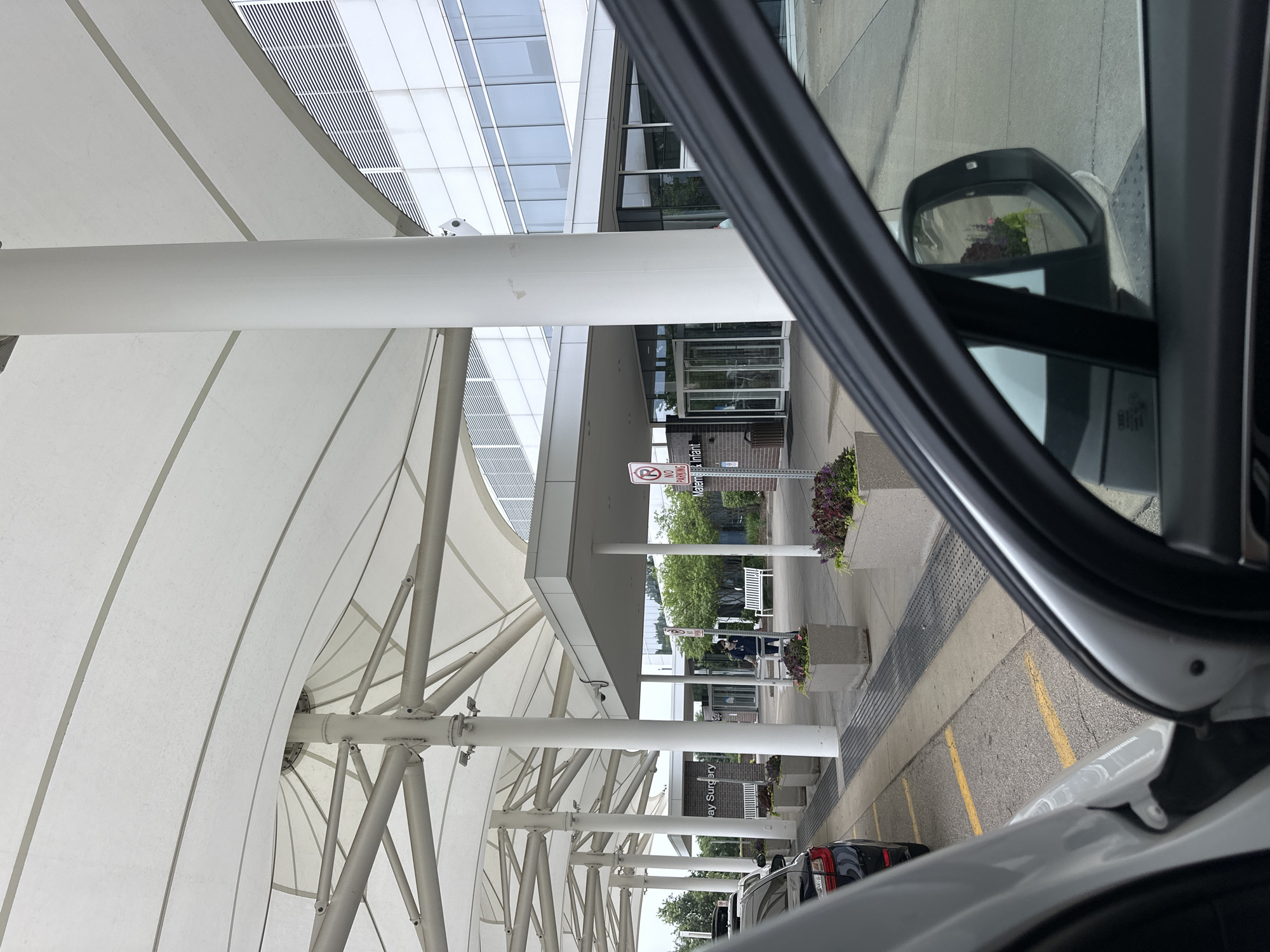 The image depicts the entrance to the "Maternity & Infant" section of a medical facility, likely a hospital, in Hoffman Estates, United States of America. The scene is captured from the perspective of someone inside a vehicle, with the car's interior and side mirror prominently in the foreground.
The building's exterior features a modern architectural style with a large, white tensile fabric canopy extending over the entrance, supported by white cylindrical pillars. Behind the canopy, a multi-story building with a facade of reflective windows and horizontal louvers is visible. The entrance itself has a covered walkway leading to revolving glass doors.
Outside the entrance, several elements are present:
* A "No Parking" sign is positioned on a pole.
* A person in a dark uniform, possibly a healthcare worker or security guard, is standing near the entrance, wearing a face mask and holding what appears to be a clipboard or tablet.
* White benches are placed along the walkway.
* Large concrete planters contain dark red and purple flowering plants.
* A black car is parked to the left of the image, with yellow lines indicating parking spaces on the asphalt ground.
* A metal grate runs along the edge of the walkway.
The overall environment suggests a functional, well-maintained public space. The lighting appears to be overcast, indicating a daytime scene. The presence of the car interior suggests the image was taken by a person arriving at or departing from the facility.