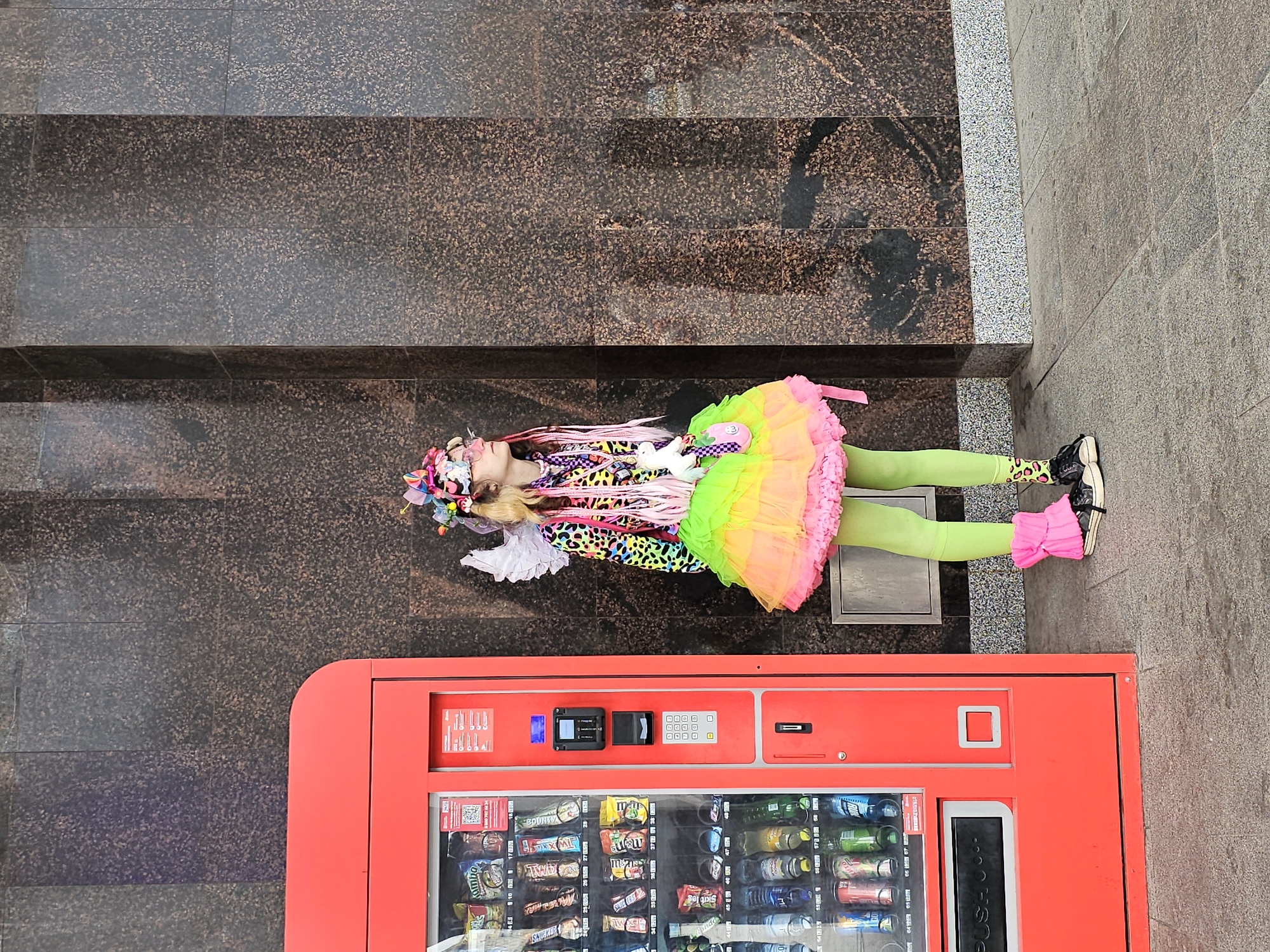 A young person in colorful, eccentric attire stands next to a bright red vending machine against a dark, textured wall. The person wears a vibrant outfit featuring a leopard-print top, a multi-colored layered skirt in shades of yellow, green, and pink, lime green leggings, and pink leg warmers over black sneakers with yellow socks. They have white fairy wings attached to their back and a festive headpiece with colorful accents. A small white unicorn plush toy is attached to their skirt. The vending machine is stocked with various snacks and drinks. The pavement in front of the vending machine and the person is made of stone slabs. The background wall is constructed of dark, speckled granite or marble tiles. The scene appears to be in an urban environment, possibly a public street or plaza in Moscow, Russia.