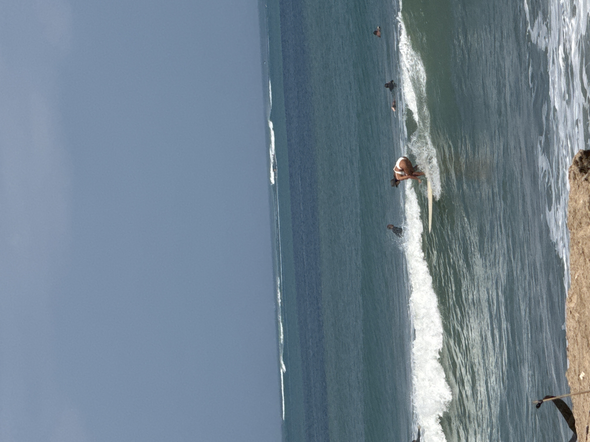 The image shows a coastline in Kuta, Indonesia, with several surfers in the ocean. The sky is overcast and the water is a deep blue-green. In the foreground, a sandy beach is visible, with some rocks and a dark object on the sand.
The main subject of the image is a female surfer riding a wave. She is wearing a white bikini and a hat, and is crouched low on her surfboard. Several other surfers are visible in the water, some of them paddling, others waiting for waves. In the background, a line of breaking waves stretches across the horizon. A boat can also be seen in the distance.