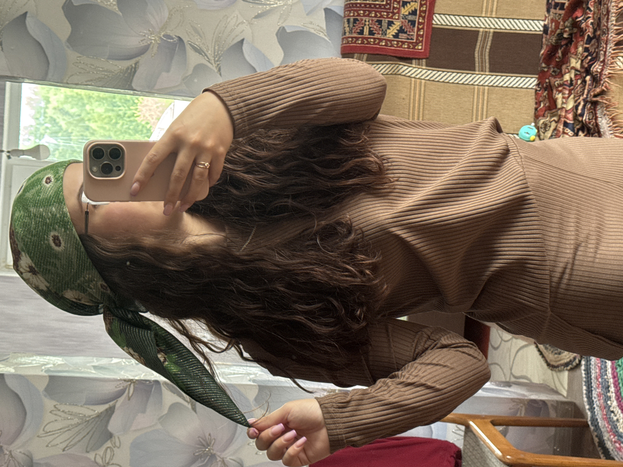 A woman is taking a selfie in a room. She is wearing a brown ribbed sweater or dress and a green patterned headscarf. Her hair is long and curly. She is holding a pink phone with multiple lenses. The background includes a window showing greenery, and wall decorations including floral patterns and rugs. The location information provided, "Nizhnyaya Gar', Russia" is not visually verifiable from the image content.