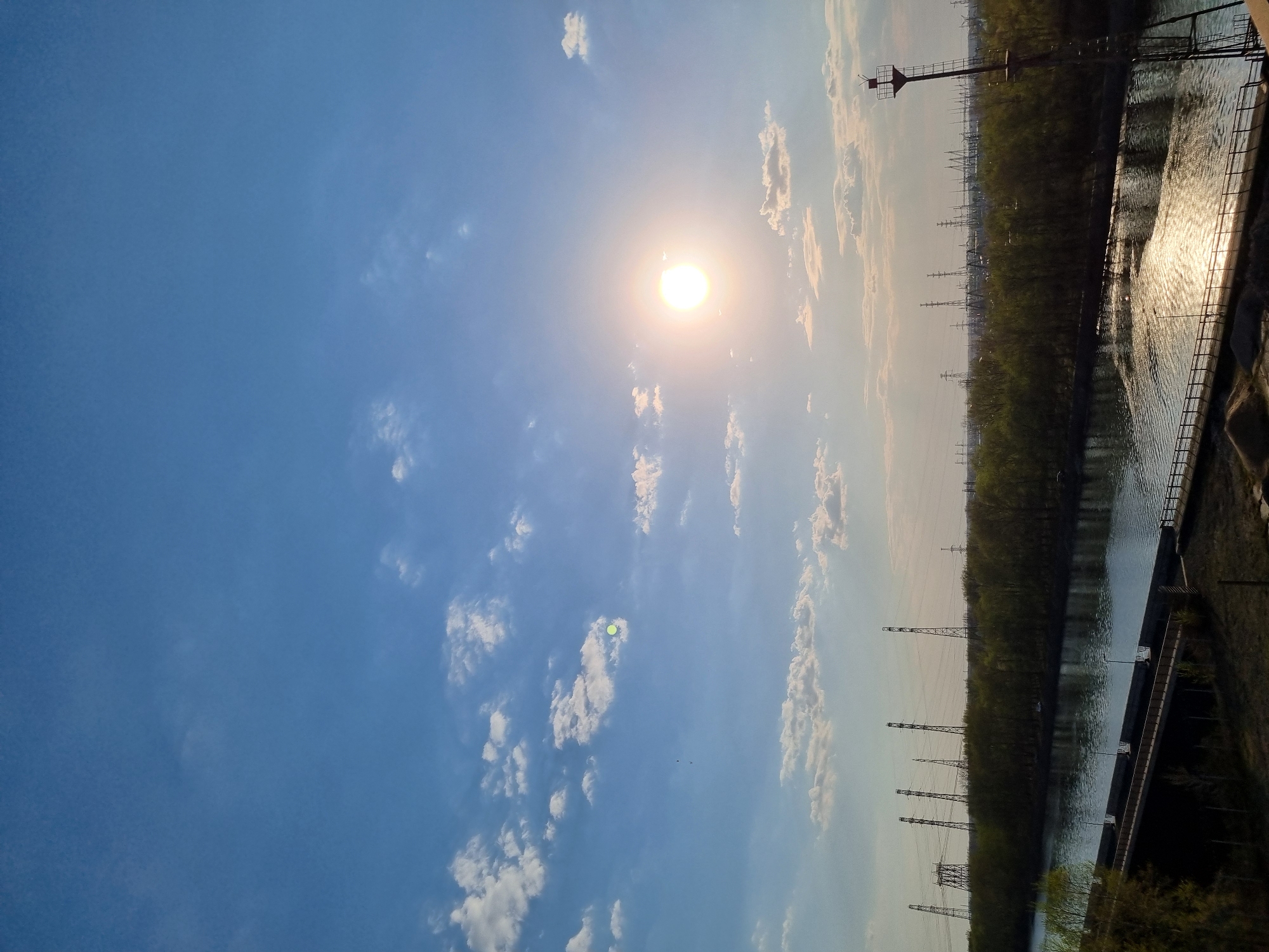 The image depicts a landscape in Chaikovsky, Russia, with a waterway reflecting sunlight on the right side. The water is bordered by a dark concrete structure and a metal railing. On the opposite side of the water is a dense line of trees stretching into the distance. Power lines and pylons are visible along the tree line, receding into the horizon. The sky dominates the left side of the image, featuring scattered white clouds and a bright sun near the center.