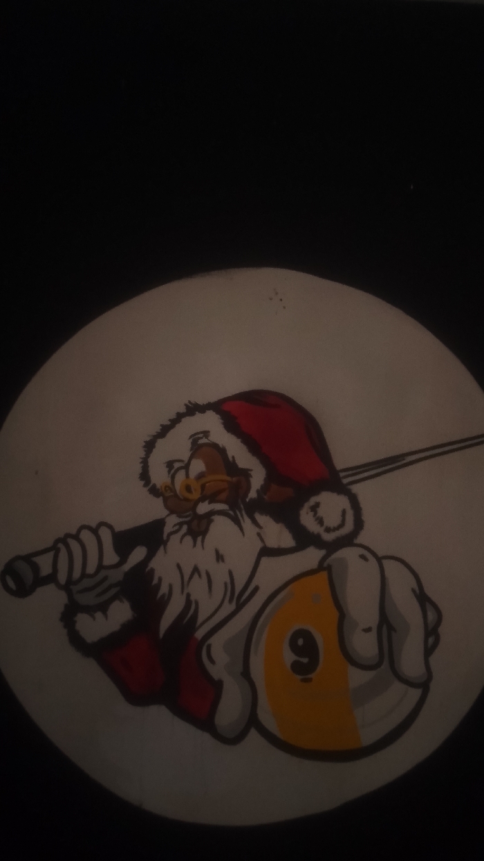 The image displays a close-up of a circular emblem or sign against a dark, featureless background. The emblem features a cartoon illustration of a brown-skinned, Santa Claus-like figure. The figure wears a red hat with white trim, yellow-rimmed glasses, and has a prominent white beard. It is depicted holding a pool cue over its right shoulder and a yellow and white striped 9-ball in its left hand. The off-white surface of the emblem shows minor surface imperfections. This scene is located in Korhogo, Côte d'Ivoire.