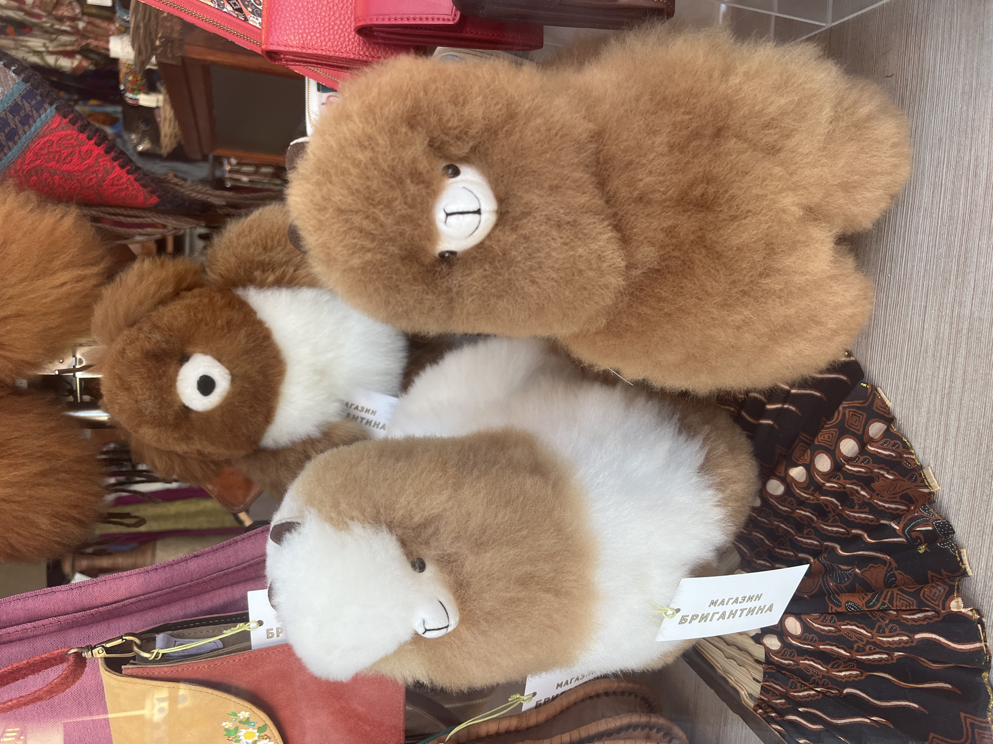 The image shows a close-up view of several stuffed animal toys displayed for sale, likely in a shop setting in Varna, Bulgaria. The toys appear to be made from alpaca wool or a similar fluffy material, giving them a soft, rounded appearance.
There are at least three prominent toy animals visible: one completely light brown, and two others with a mixed brown and white coloration. Each toy has basic facial features, including eyes and a simple mouth. The toys are arranged somewhat haphazardly, with some partially obscuring others.
Several other items are visible in the background. There are purses, wallets, and other accessories, adding to the impression that this is a retail environment. One white tag is attached to a toy with text in Russian, including the word "Бригантина" (Brigantina), which may be the name of the shop.
The backdrop consists of neutral surfaces, including what looks like a wooden counter or shelf, which is possibly made from bamboo. The composition suggests a display meant to entice shoppers in the Bulgarian seaside city.