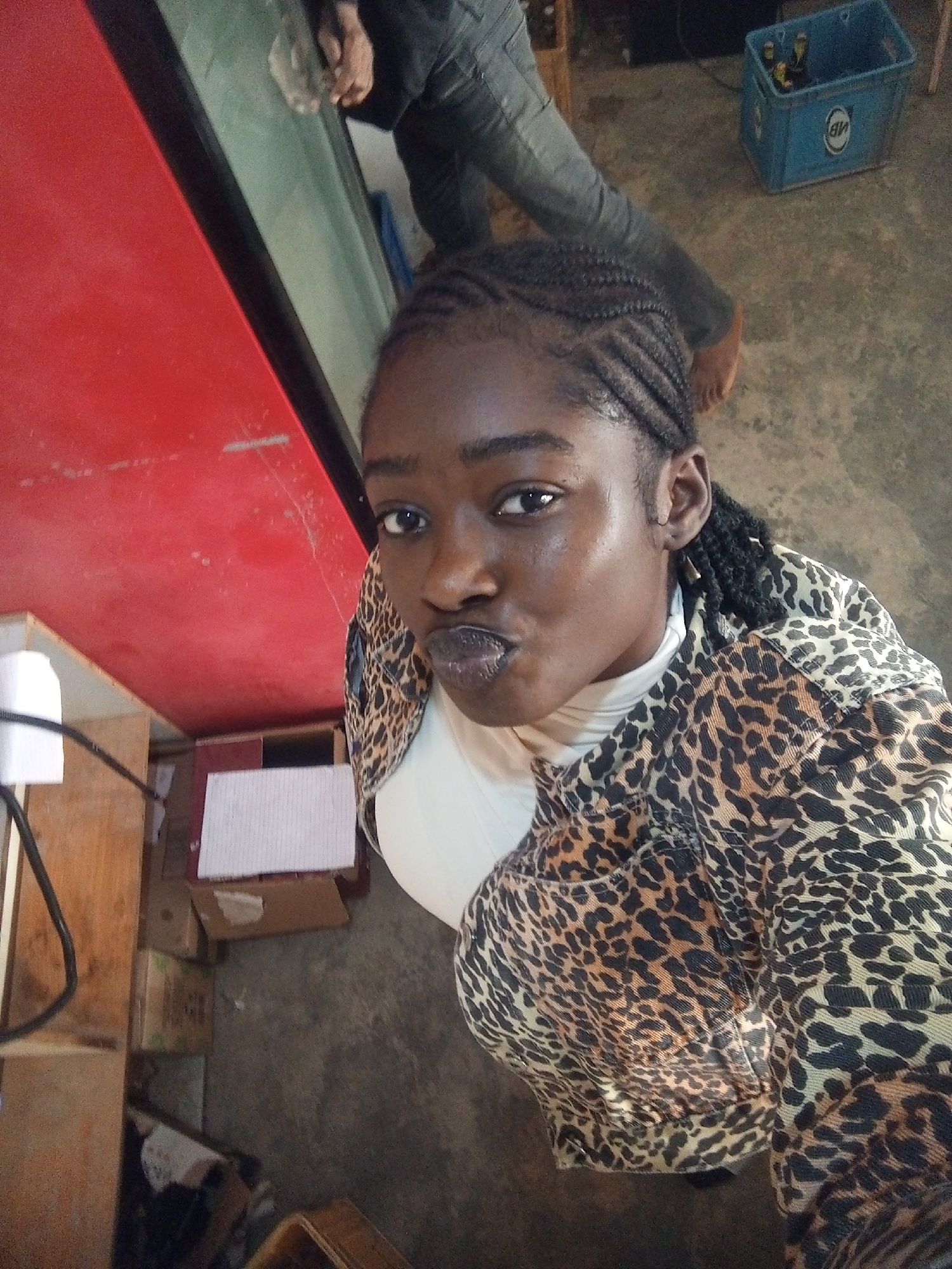 The image captures a close-up, angled view of a young Black woman with braided hair, wearing a white turtleneck and a leopard-print jacket. She is looking directly at the camera with her lips pursed. In the background, to the left, a portion of a red surface is visible, possibly a refrigerator door or wall. To the right, a blue plastic crate filled with bottles, likely beer, rests on a concrete floor. Further back, another person's legs, clad in dark pants, are partially visible, with one bare foot on the floor. The environment appears to be indoors, with a rustic or utilitarian feel, suggested by the concrete floor and visible shelving units with boxes. The lighting is somewhat dim and uneven.