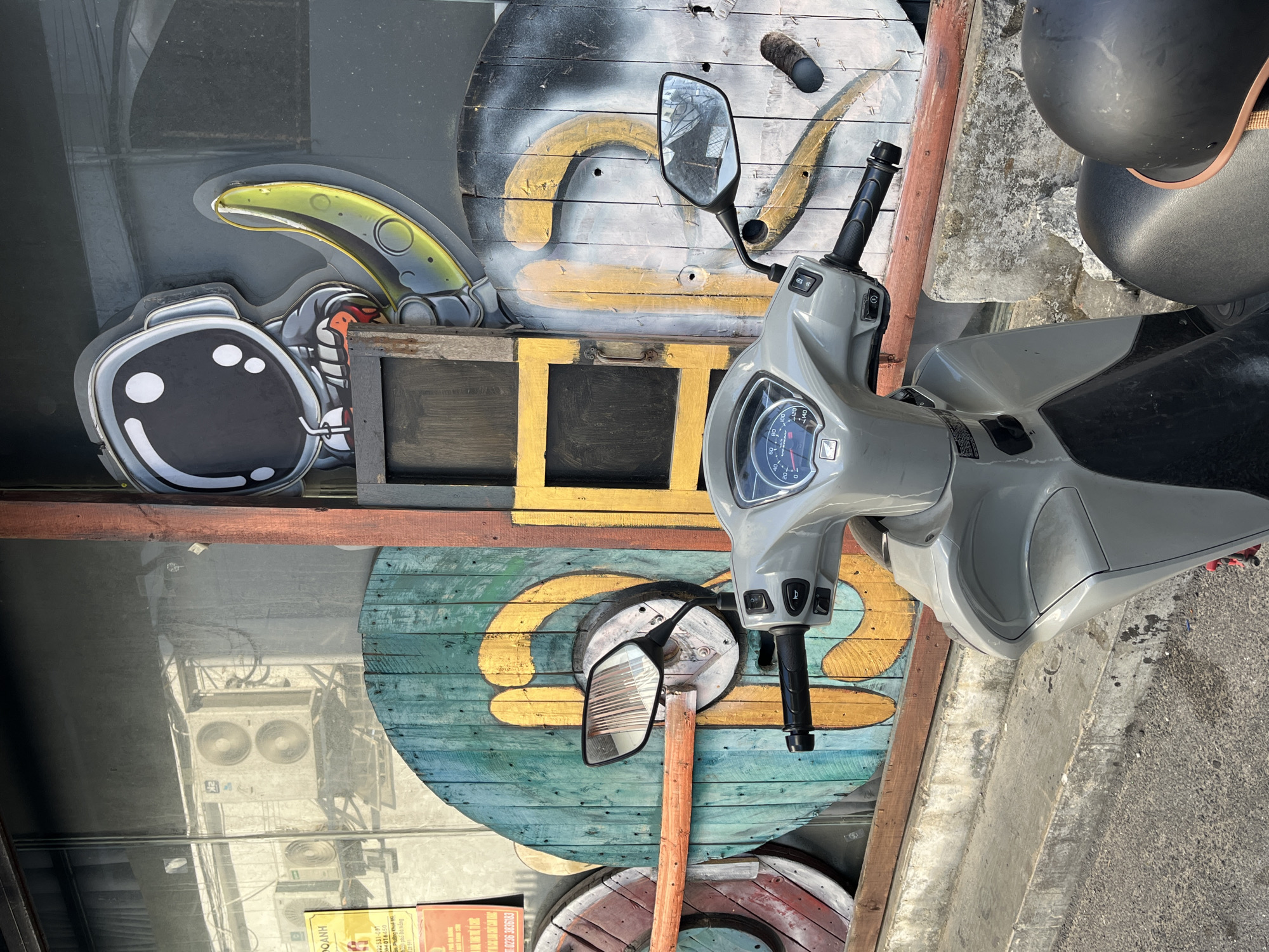 A grey scooter is parked next to a wall decorated with large, painted wooden cable spools. The spools feature abstract, swirl-like designs in yellow, white, and teal. A cartoon astronaut figure is mounted to the wall behind the scooter. The scooter is parked on a concrete surface next to the wall. The image is taken from an eye-level perspective. Reflected in the window, behind the astronaut figure, are air conditioning units.