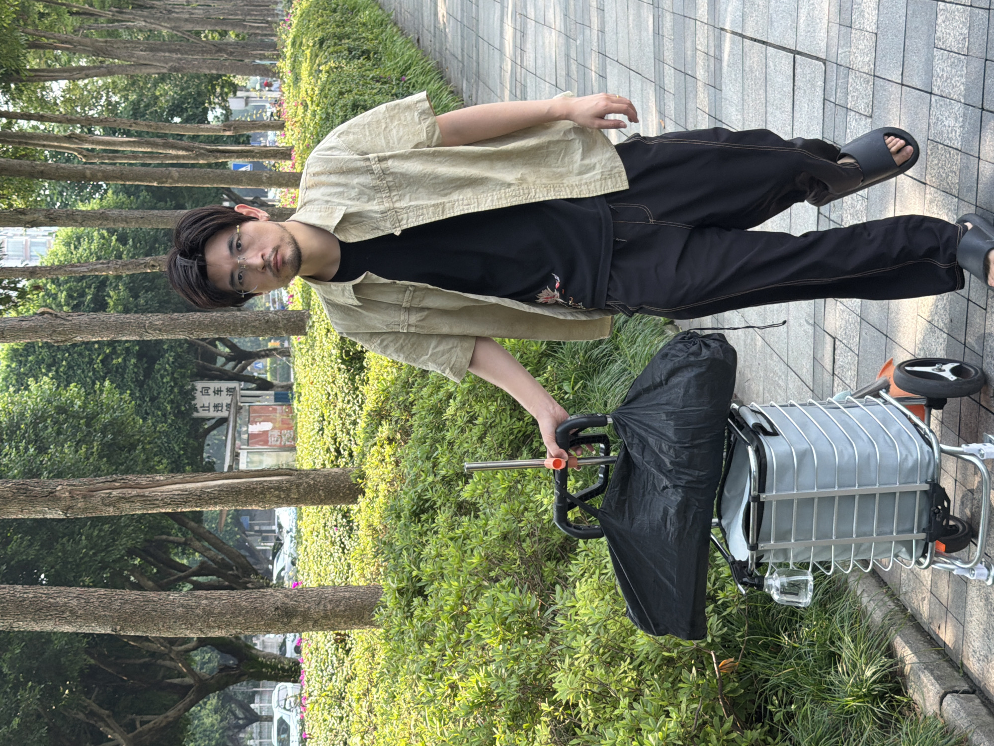 A young East Asian man with glasses, a beard, and dark hair is walking on a paved sidewalk lined with trees and lush green bushes. He is wearing a light-colored, short-sleeved collared shirt over a black t-shirt, black pants, and black slides. He is holding the handle of a light gray shopping cart with a black bag on top. The cart has two large wheels at the back and two smaller wheels at the front. In the background, there are more trees, some buildings, and parked cars. A street sign with Chinese characters is visible on the left side of the image. The scene appears to be an urban park or a tree-lined street in the city of Jiangjin, China. The man is looking forward, and his body is angled slightly towards the camera.