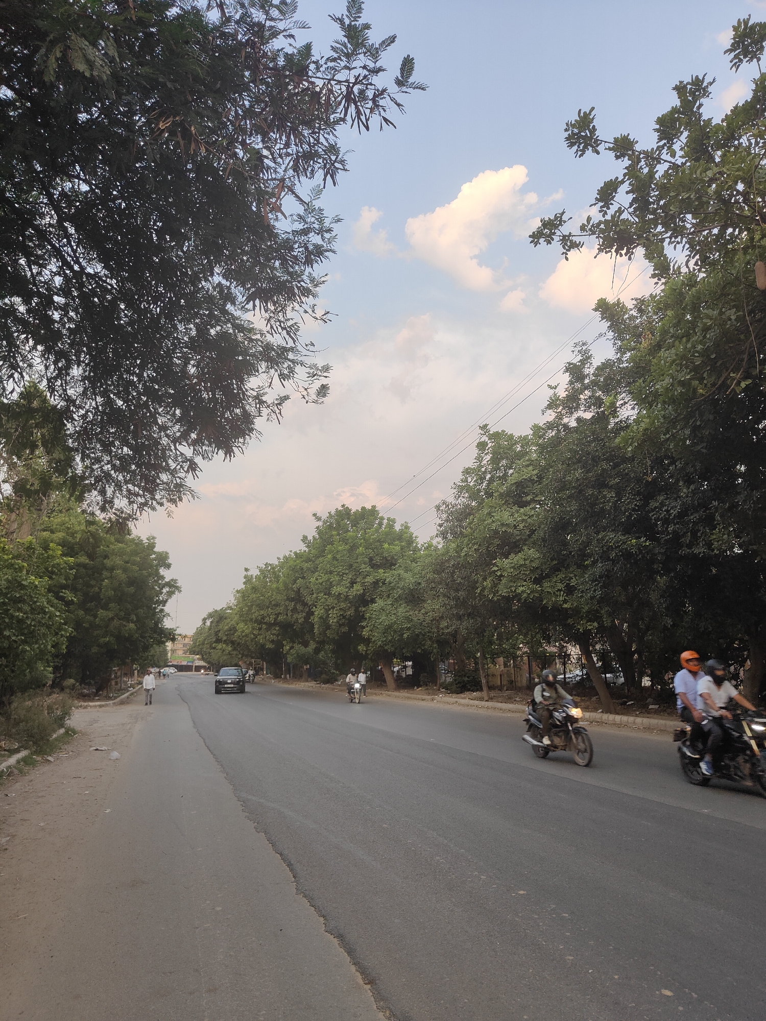 The image shows a street scene in Gurgaon, India, with lush green trees lining both sides of an asphalt road. The sky is partly cloudy, with soft, diffused sunlight. On the right side of the road, two men are riding a motorcycle, with the rider in front wearing a blue shirt and an orange helmet, and the passenger wearing a white shirt. Another motorcycle is seen further ahead on the right lane, with its headlight on. A black car is driving in the left lane, and a single person is walking on the sandy shoulder on the left side of the road. The environment appears to be an urban or suburban area with well-maintained trees providing shade.