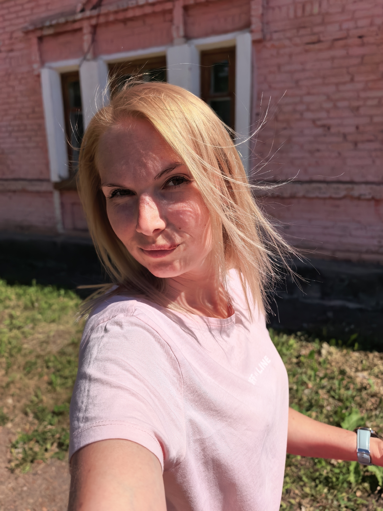 A woman with blond hair is posing outdoors in front of a pink brick building with white-framed windows. She is wearing a light pink t-shirt. There is grass and patchy ground visible in the foreground. The location is assumed to be in Sterlitamak, Russia.