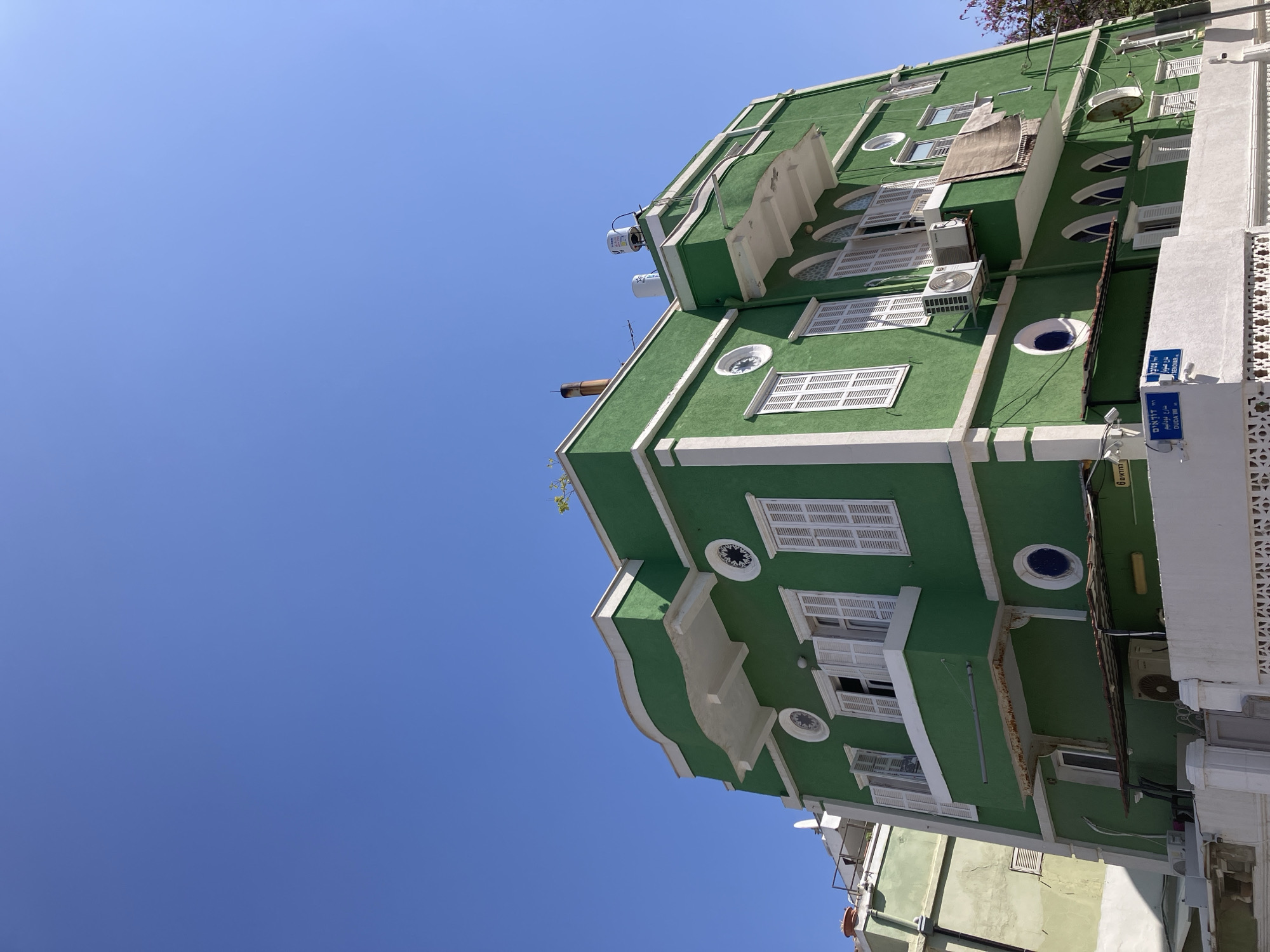 The image captures a building located in Tel-Aviv, Israel, standing out with its green facade and white trim. The building features a multi-story structure with a mix of rectangular and circular windows, some adorned with white shutters. Several air conditioning units are mounted on the exterior walls. A street sign, with text in Hebrew and English, indicates the street name "DAUDA I.M. GEISS." The sky overhead is a clear, unblemished blue.