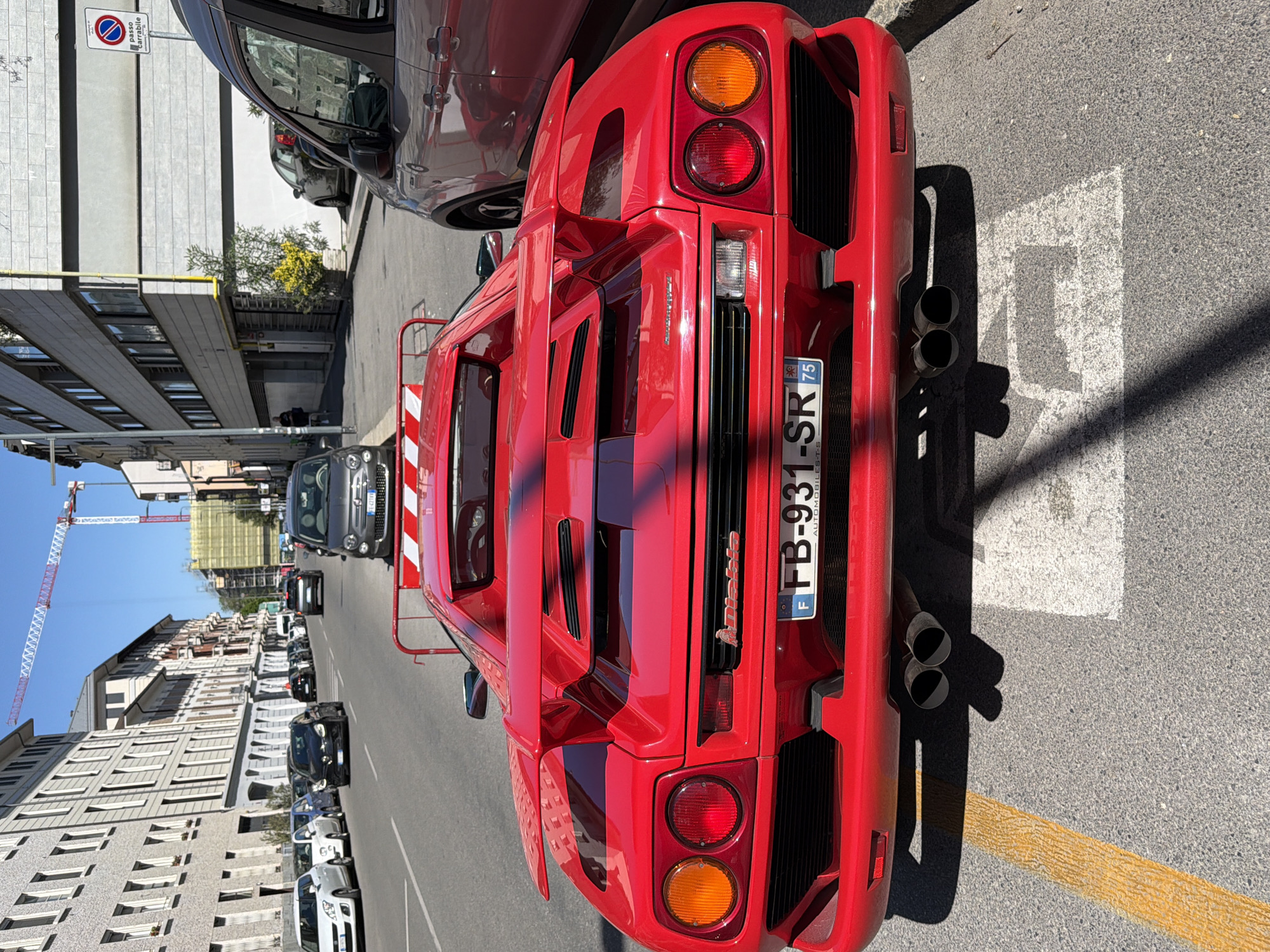 The primary subject of this image is a vibrant red Lamborghini Diablo supercar, prominently parked on an urban street in Milan, Italy. The image is oriented vertically, showcasing the car's rear view with the street extending horizontally across the frame.
The supercar exhibits its distinctive design, featuring four circular taillights (two red and two amber), a prominent rear spoiler, and multiple vented panels on the engine cover. Four large, dark exhaust pipes are visible on the right side of the rear bumper. The license plate, "FB-931-SR 75," includes a French "F" oval and the text "AUTOMOBILES," indicating French registration. The car's model name, "Diablo," is also clearly badged on the rear.
The street is bordered by multi-story buildings, some displaying classic architectural elements, others more contemporary. Numerous other vehicles, including various sedans and hatchbacks, are parked along the curb or seen in the distance. A "Passo Carrabile" (driveway access, no parking) sign, marked with a red circle and blue slash symbol, stands on a pole adjacent to the red car. Road markings, such as a white outlined parking space and a yellow no-parking line, are visible on the asphalt. In the background, a large construction crane and scaffolding suggest ongoing building activity under a clear blue sky.
The scene is brightly lit by strong daylight, casting defined shadows from the vehicles and buildings, indicating it is mid-day. No people are visibly engaged in any specific activities.