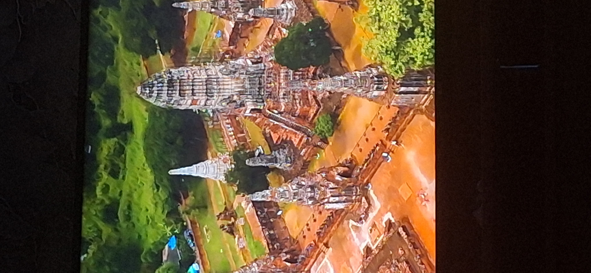 The image is an aerial view of an ancient temple complex with multiple stupas and ruins. The architecture suggests Southeast Asian influence, likely Buddhist. The central and largest structure is a tall, multi-tiered tower with intricate carvings, surrounded by smaller spires and brick structures. The complex is situated in a lush green landscape with trees and foliage. The ground within the complex is a reddish-brown, possibly mud or weathered stone. There are a few small figures visible, appearing as people walking around the site. In the background, there are hints of modern structures, including blue tarps and what appear to be vehicles, suggesting proximity to a populated area.
This image does not depict the city of Bila Tserkva, Ukraine. The architectural style and landscape are inconsistent with the geographical and cultural context of Ukraine.