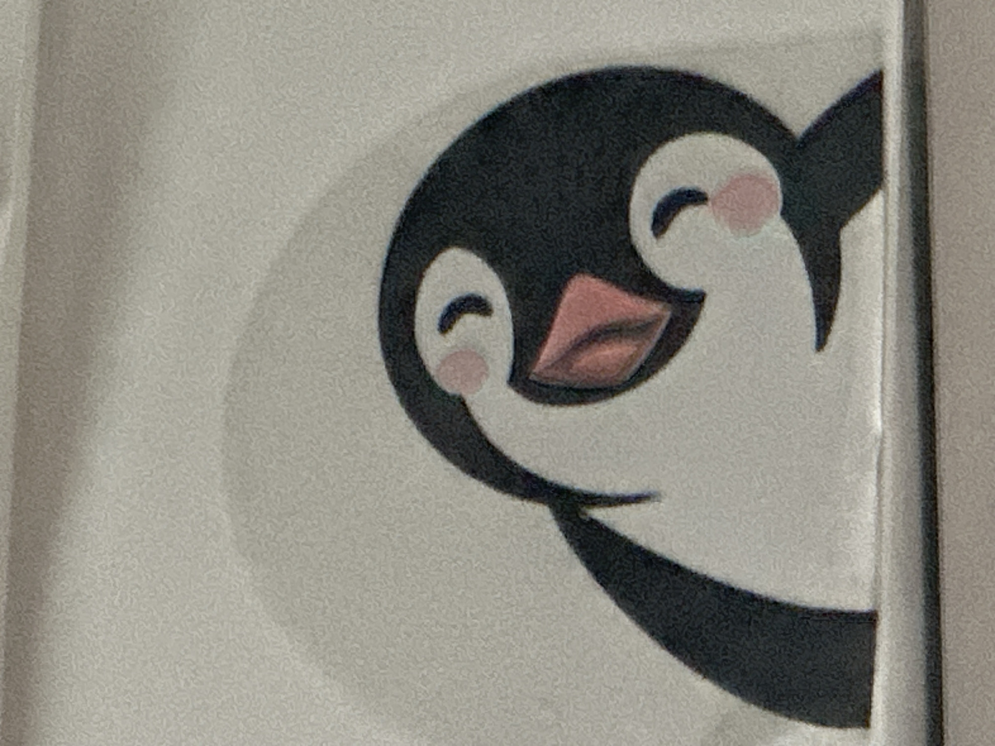 The image shows a close-up of a stylized illustration of two penguins. The larger penguin's head and upper body are visible, with its beak open and a pink inner mouth. A smaller penguin is nestled against its chest, with its head tilted up towards the larger penguin's beak. Both penguins have black bodies, white faces, and pink cheeks. The background is a light, textured gray, with a subtle shadow cast on the upper left. A dark horizontal line runs across the bottom of the image, suggesting a surface or edge. There is no discernible location in the city of Chaikovsky, Russia, visible in this image.