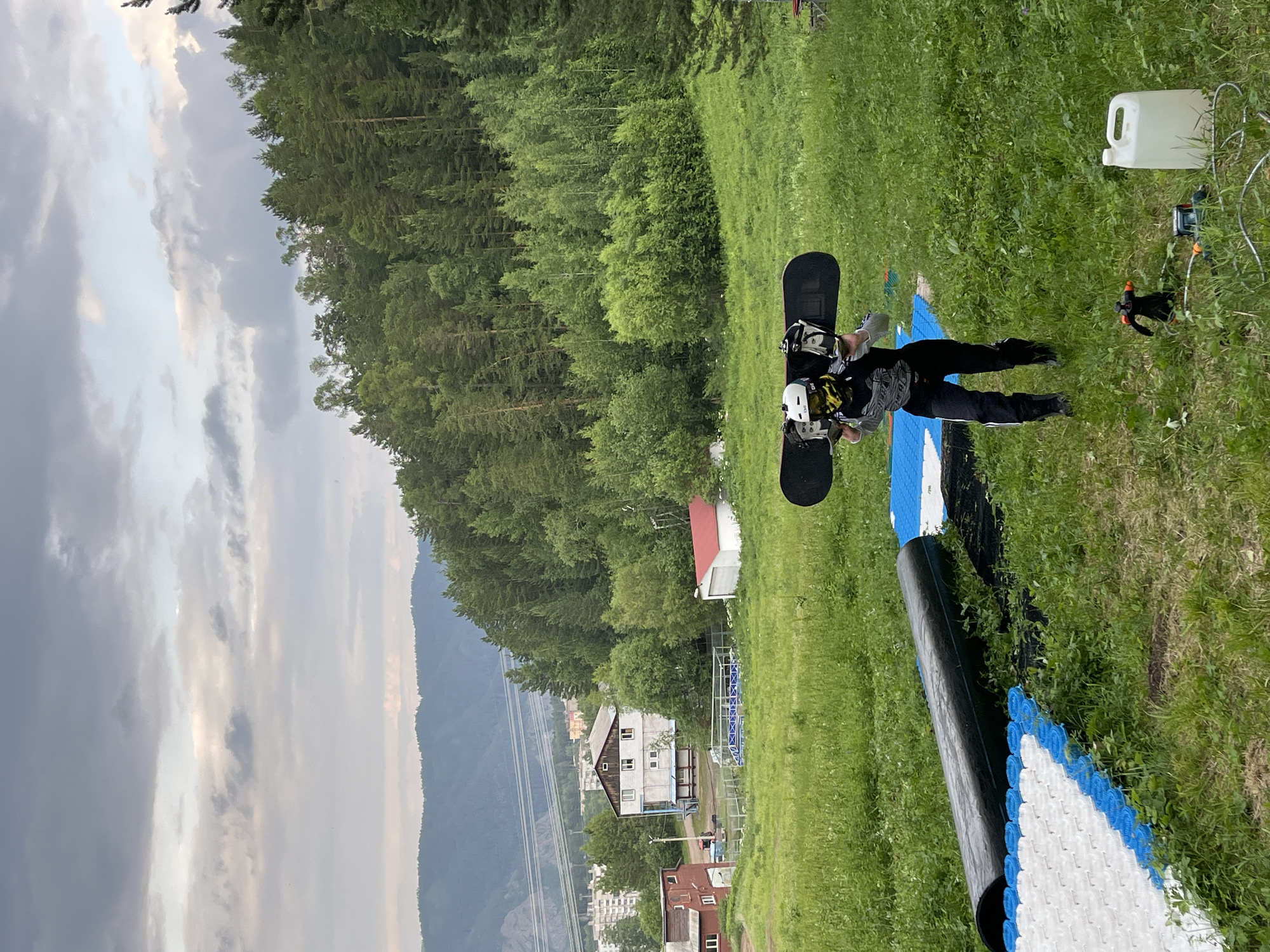 A person lies on the ground, on their back, with a snowboard attached to their feet. They are wearing a helmet, goggles, and dark-colored snowboarding attire. The person appears to be in a grassy area. To their right, there is a white plastic container and some equipment, possibly for snowmaking or maintenance. In the background, there is a dense forest and a mountain range. The sky is cloudy, with a mix of gray and white clouds. The lighting suggests it is daytime, possibly late afternoon.
The scene is set in a mountainous, forested area, with buildings and what appears to be a ski lift infrastructure visible in the distance, suggesting a ski resort or a similar recreational area. The presence of the snowboard and the person's attire indicate a snowboarding context, even though the ground is covered in grass, implying it's either the off-season or a summer training setup. The person's pose and the equipment around them do not convey any specific emotion, appearing to be a static scene. No visible text is present in the image. The location is identified as Divnogorsk, Russia.