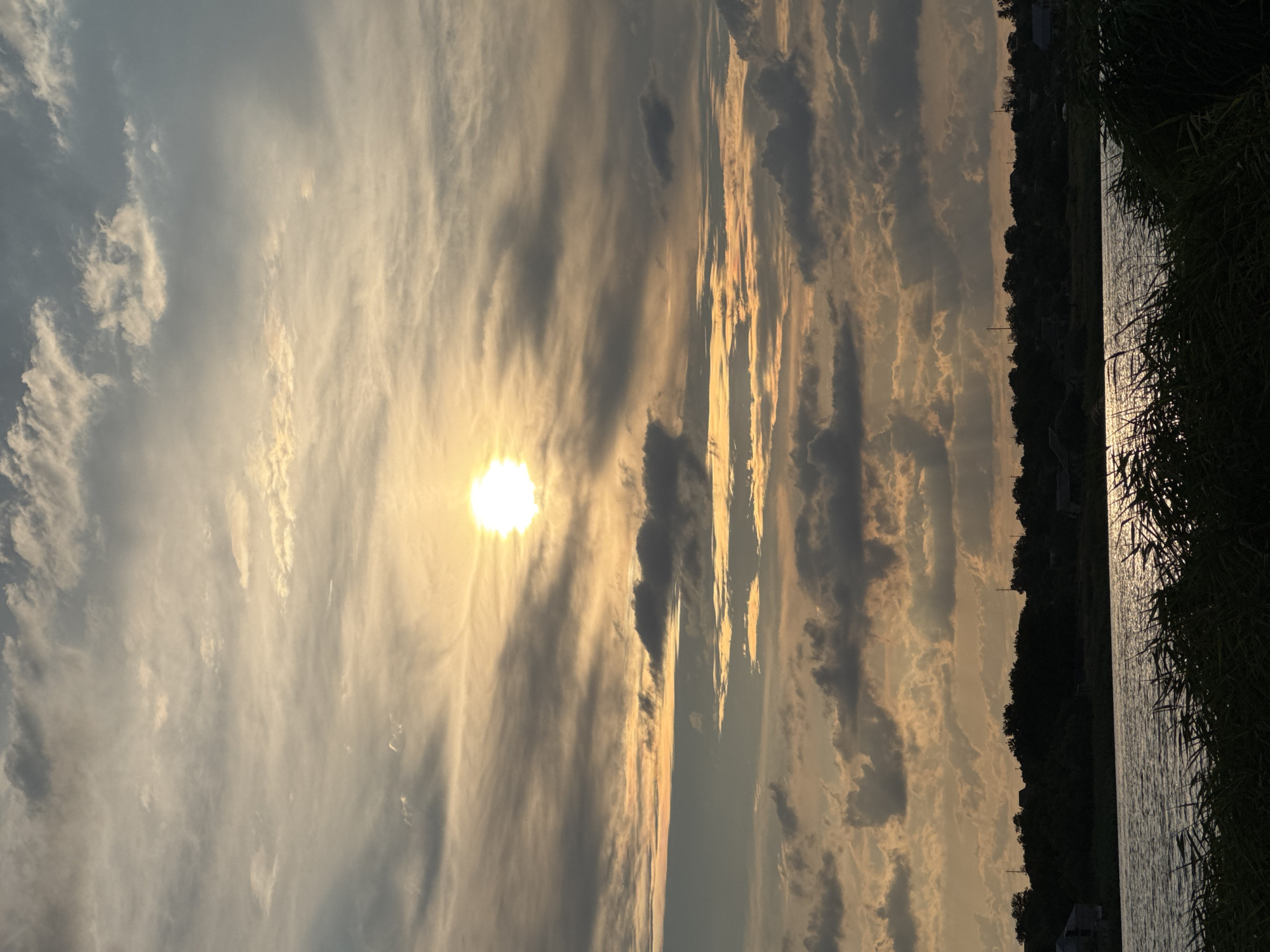 The image depicts a sunset over a body of water, likely a river or lake, in Karachunivka, Ukraine. The sun, a bright orb, is partially obscured by clouds, casting a warm, golden light across the sky. The clouds are varied in texture and color, ranging from thin, wispy cirrus formations to darker, more ominous cumulus clouds. Below the sky, a dark silhouette of trees and distant buildings lines the horizon, suggesting a rural or semi-rural setting. In the foreground, the surface of the water reflects the golden hues of the sunset, with streaks of light shimmering on its surface. Tall grasses or reeds are visible along the edge of the water, their dark forms silhouetted against the illuminated water and sky. The overall impression is one of natural beauty and tranquility as day transitions into evening.