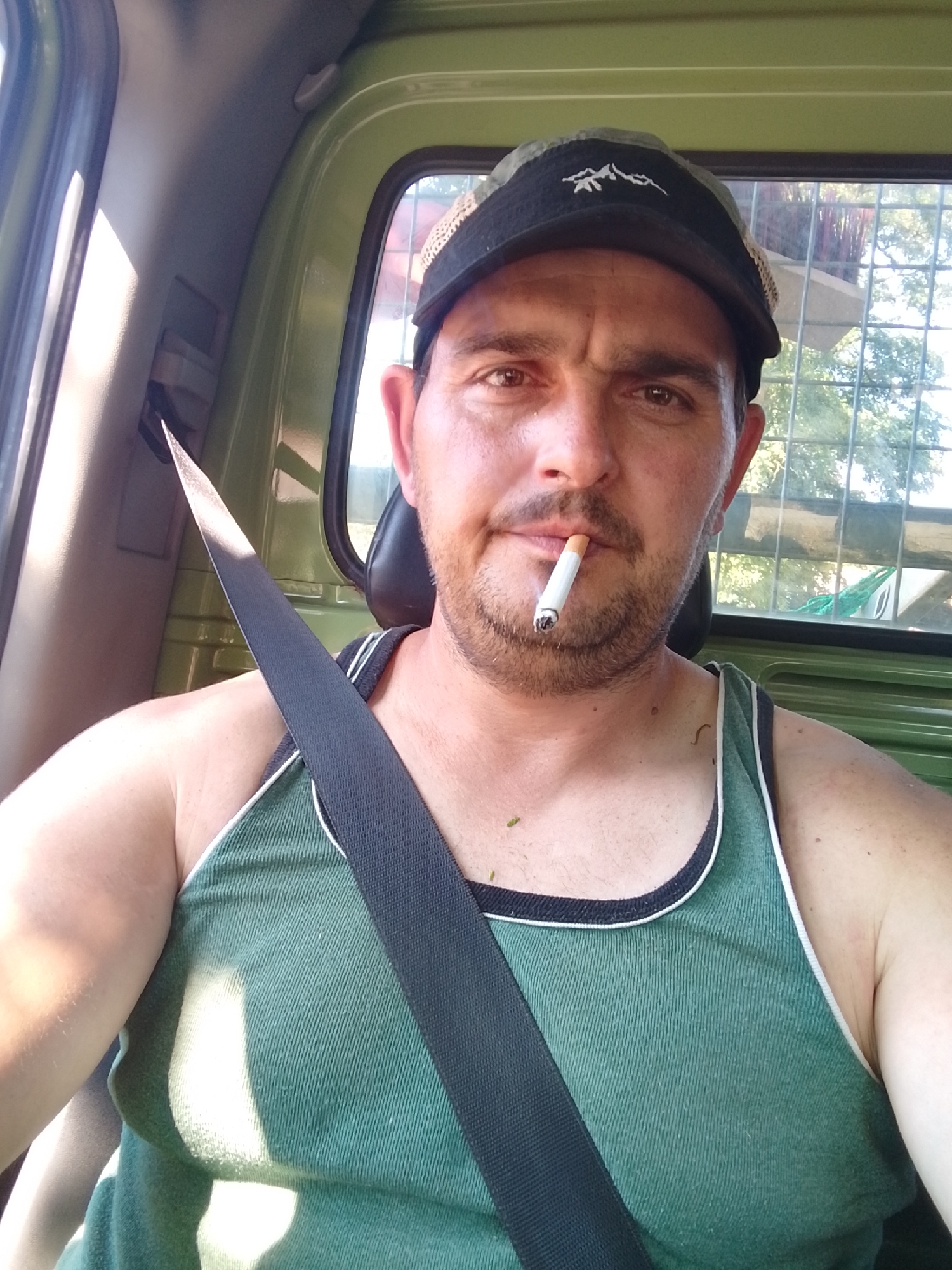 A man is sitting in the driver's seat of a green vehicle, wearing a green tank top, a black cap with a mountain logo, and a black seatbelt across his chest. He is smoking a cigarette. The interior of the vehicle appears to be a truck cab. The background shows foliage and a fence, suggesting an outdoor environment. The time of day appears to be daytime, with sunlight casting shadows. There is no identifiable information about the location being in Wedel, Germany.