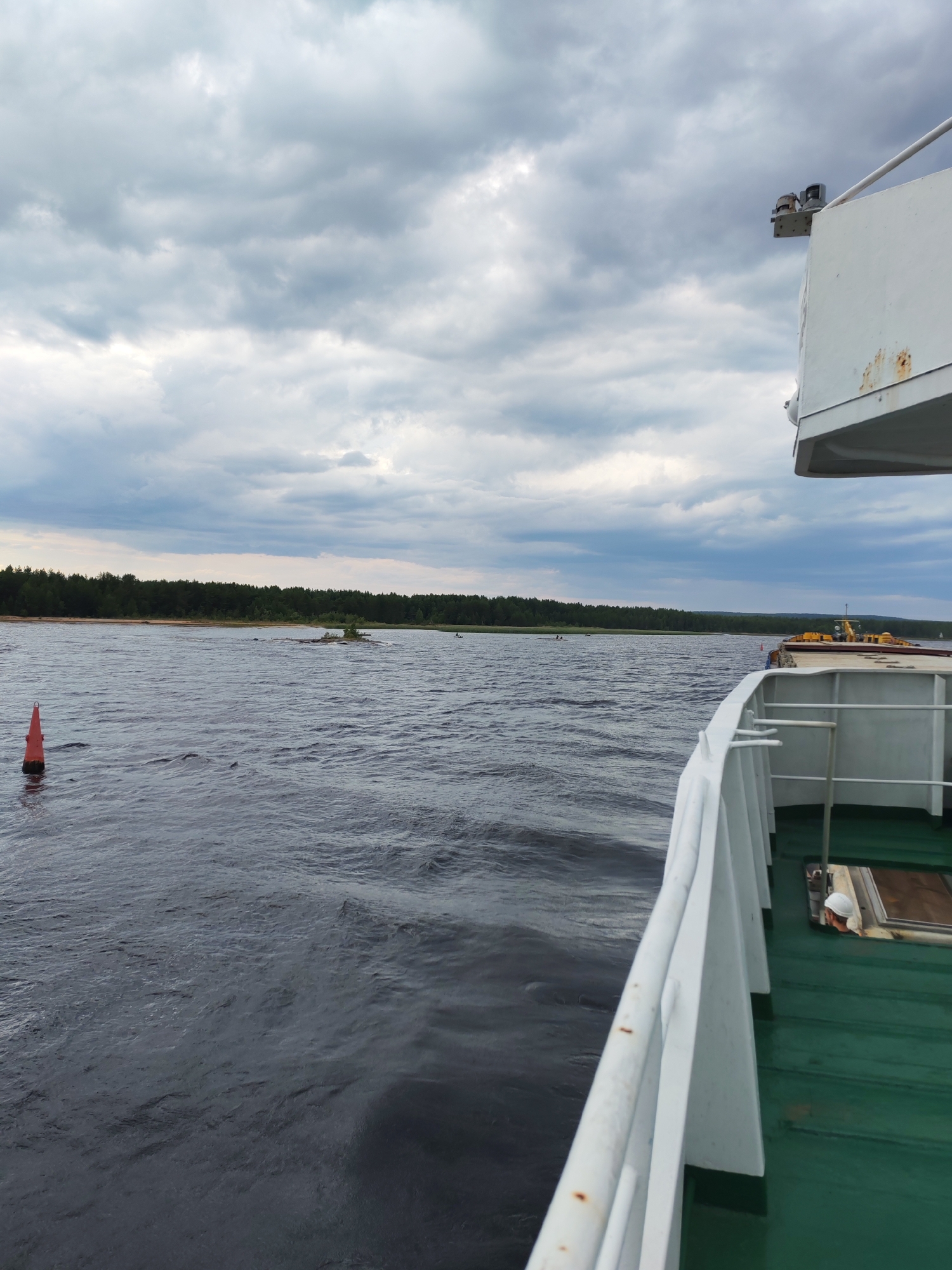 A forward-facing, eye-level, medium shot captures a body of water from the deck of a ship. The water is dark and choppy, with small waves rippling across its surface. In the distance, a dense, dark green tree line borders the horizon, with a strip of lighter sand or beach visible just before the trees. A small, rocky island with sparse vegetation sits in the middle of the water, slightly to the left of center. A single, red, conical buoy is positioned in the foreground, on the left side of the frame, partially submerged in the water.
The right side of the image shows the deck and railing of the ship from which the photo is taken. The deck is painted green, and a white railing runs along the edge. A portion of the ship's superstructure is visible at the top right, with some signs of rust and weathering. A person wearing a white hard hat is partially visible on the green deck, looking downwards. The sky above is heavily overcast with thick, gray clouds, suggesting a cloudy or potentially stormy day. The scene appears to be set in a natural, possibly remote, waterway, consistent with the location of Shalsky, Russia, known for its river and forest landscapes.