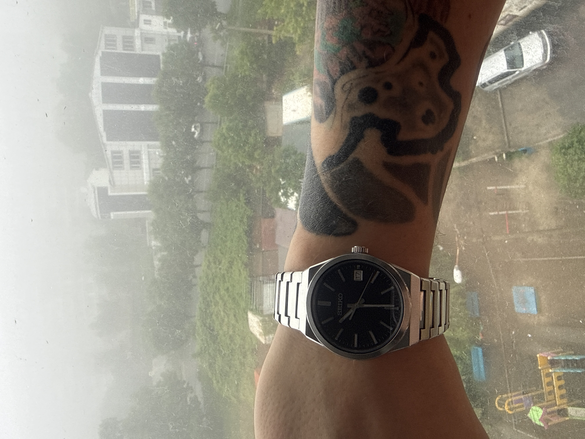 The image is a first-person perspective photograph, taken indoors through a dirty window. A person's left arm, adorned with a detailed black tattoo, is prominent in the foreground, wearing a silver Seiko wristwatch with a black dial. The watch displays the time as approximately 10:17, with the date window showing '17'.
The background, visible through the smudged window, depicts a hazy, overcast day with rain. In the middle ground, there are rows of trees and several multi-story buildings, some appearing to be apartment complexes. The architectural style suggests a post-Soviet era.
In the lower part of the image, a playground with colorful structures is visible, along with a silver car parked on a paved area. The overall environment appears to be an urban residential area. The presence of the buildings, playground, and parked car, along with the general urban landscape, strongly indicates a location within the city of Vladivostok, Russia. The weather conditions suggest it is likely a mild to warm season, given the greenery.