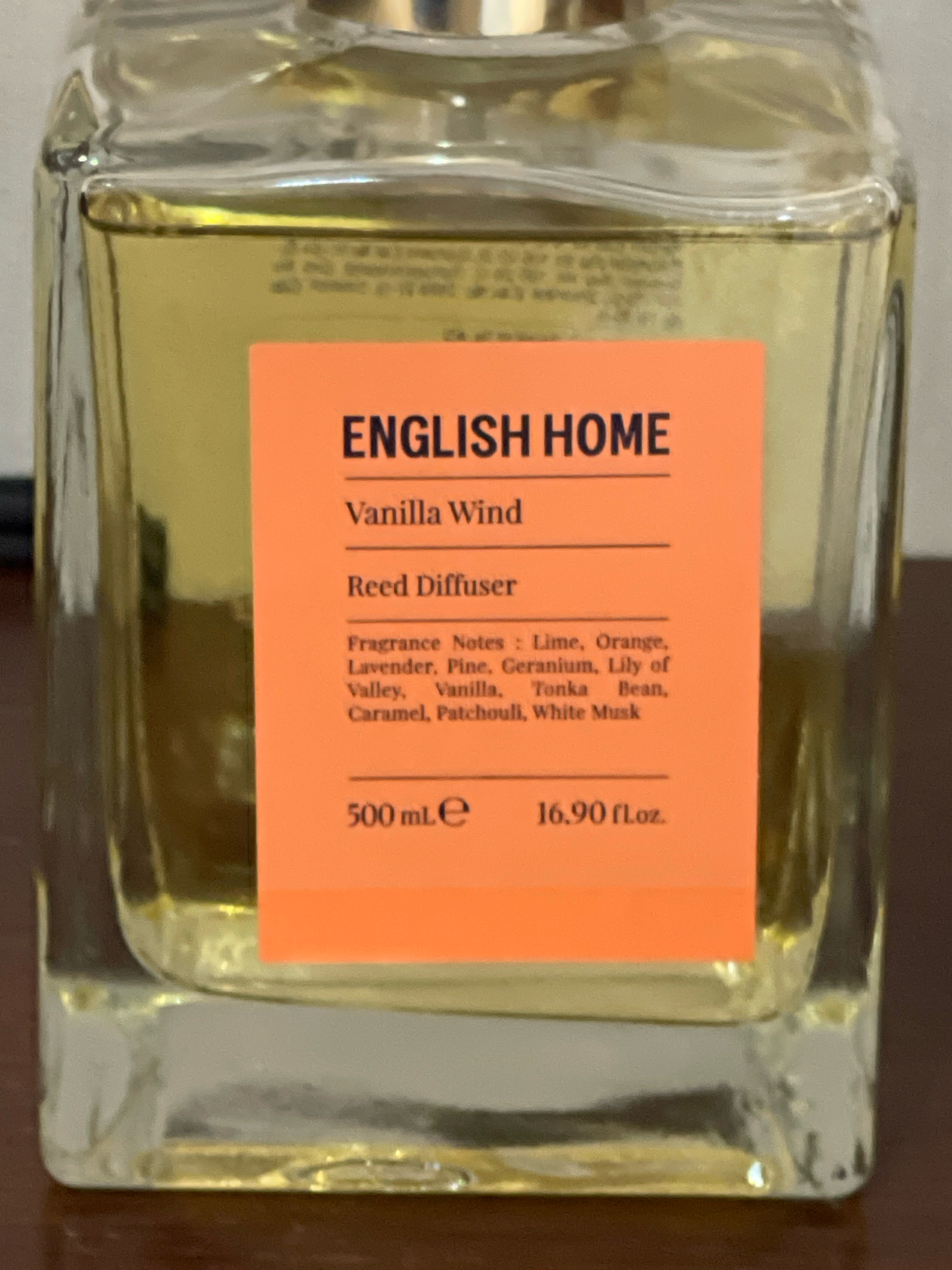 A clear glass perfume bottle, partially filled with a yellow liquid, sits on a brown wooden surface. An orange label is affixed to the front of the bottle. The label contains text in black. At the top of the label, the words "ENGLISH HOME" are printed. Below this, "Vanilla Wind" is written, followed by "Reed Diffuser". Further down, a list of "Fragrance Notes" is provided: "Lime, Orange, Lavender, Pine, Geranium, Lily of Valley, Vanilla, Tonka Bean, Caramel, Patchouli, White Musk". At the bottom of the label, "500 mL e" is printed to the left, and "16.90 fl.oz." to the right. The background is blurred, but appears to be a domestic indoor setting. There are no people visible in the image. The location is Jabriya, Kuwait.