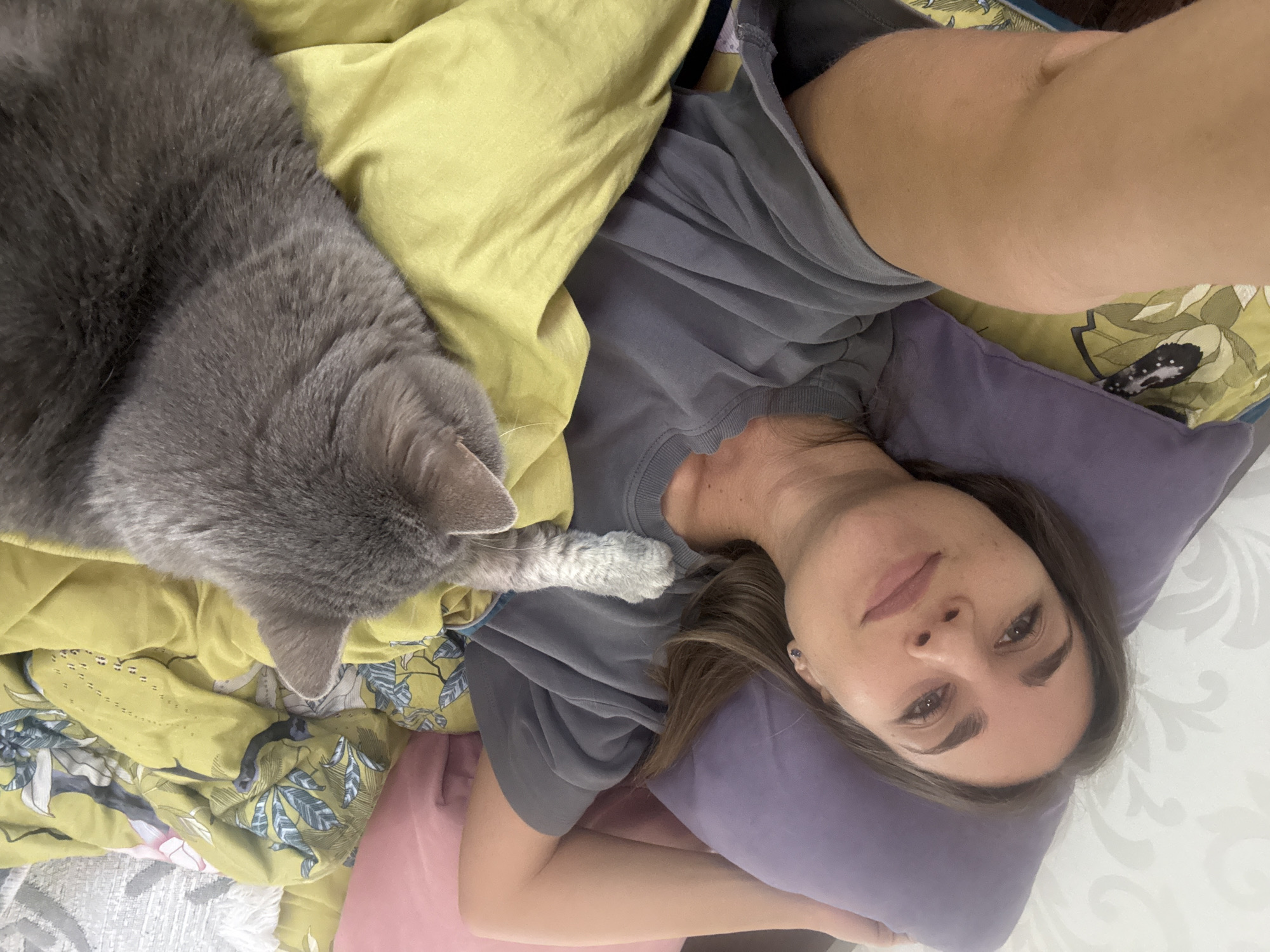 A woman lies on her back on a bed covered in floral-patterned bedding and a yellow blanket. She is using a purple pillow. She is wearing a grey T-shirt. A grey cat is lying to her left, its paw resting on her shirt. The location is Zanevka, Russia.