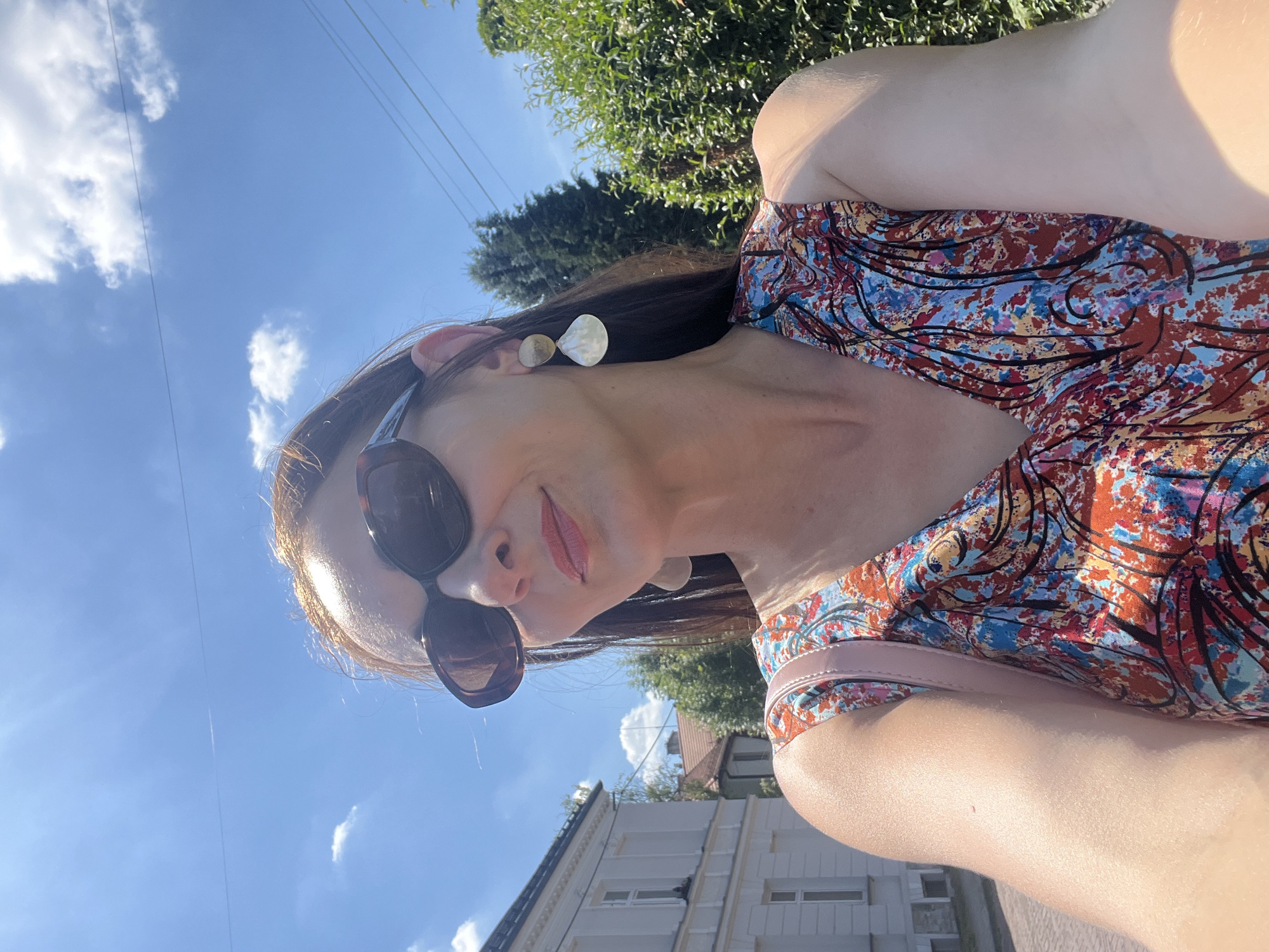 The image shows a woman taking a selfie outdoors. She is wearing a colorful, patterned dress and large sunglasses. The background is a bright blue sky with some white clouds. There are trees and buildings visible in the distance, suggesting an urban or suburban setting. The lighting indicates it is daytime, likely a sunny day. The woman's face is partially visible, and she has a slight smile. She is also wearing large, pale earrings. A pink strap from what appears to be a bag is visible across her shoulder. The image is oriented vertically, with the top of the frame showing the sky and the bottom showing buildings and greenery.