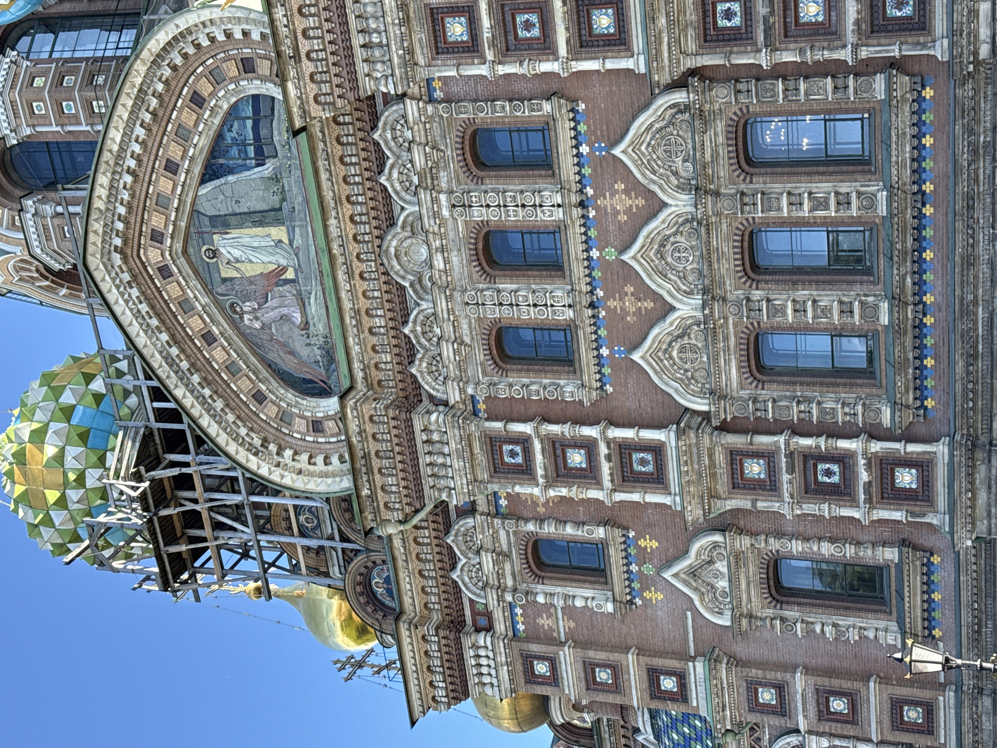 This image shows the exterior of the Church of the Savior on Spilled Blood in Saint Petersburg, Russia. The building's ornate facade is detailed with intricate brickwork, colorful mosaic tiles, and decorative stonework surrounding arched windows. Several of these windows are visible, reflecting the bright blue sky. One large, arched mosaic depicting religious figures is prominently featured. A colorful, multi-faceted dome, likely one of the church's signature onion domes, is partially visible on the left, supported by scaffolding. A smaller golden dome and a cross-topped spire are also visible against the clear blue sky. The bright sunlight suggests it is daytime. The overall impression is one of rich architectural detail and religious significance.